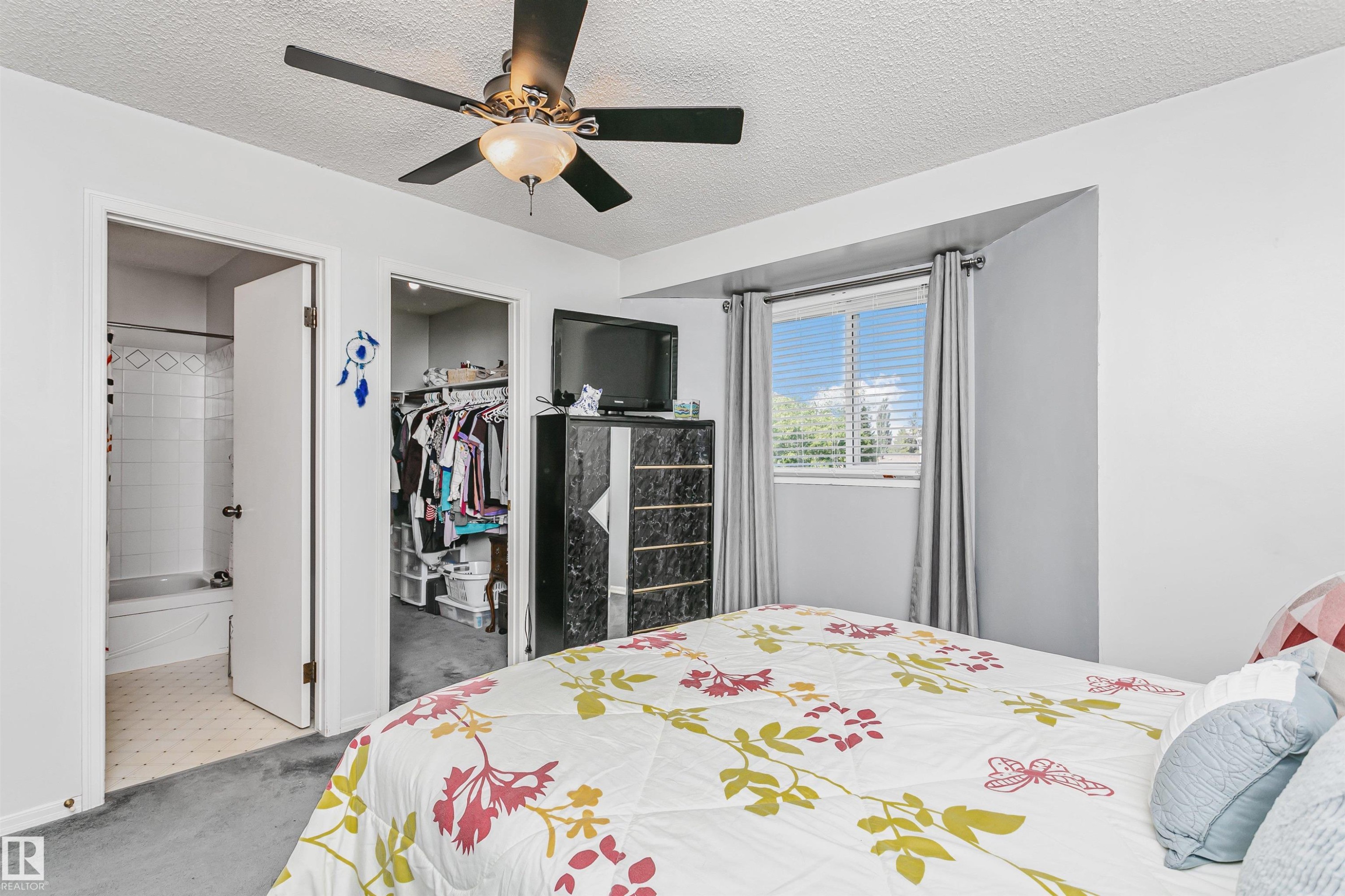 Interior room featuring a window with horizontal blinds, a ceiling fan with light fixture, gray carpeting, a walk-in closet, and a white tiled bathroom with a bathtub - 1612 80A Street, Edmonton, AB