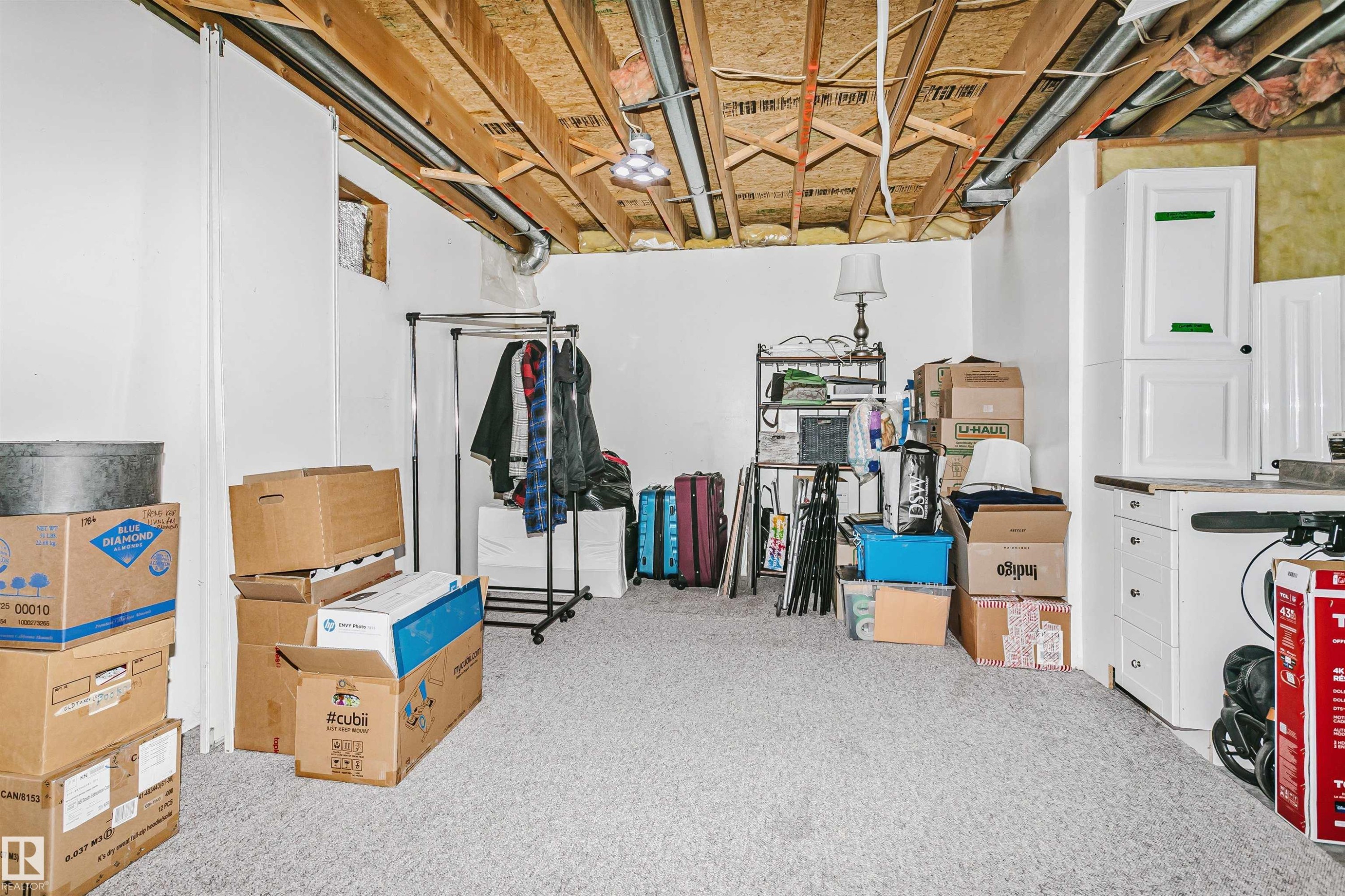 Unfinished basement space featuring exposed wood joists and ductwork, white painted walls, light grey carpeting, and built-in white cabinetry with a countertop - 1612 80A Street, Edmonton, AB