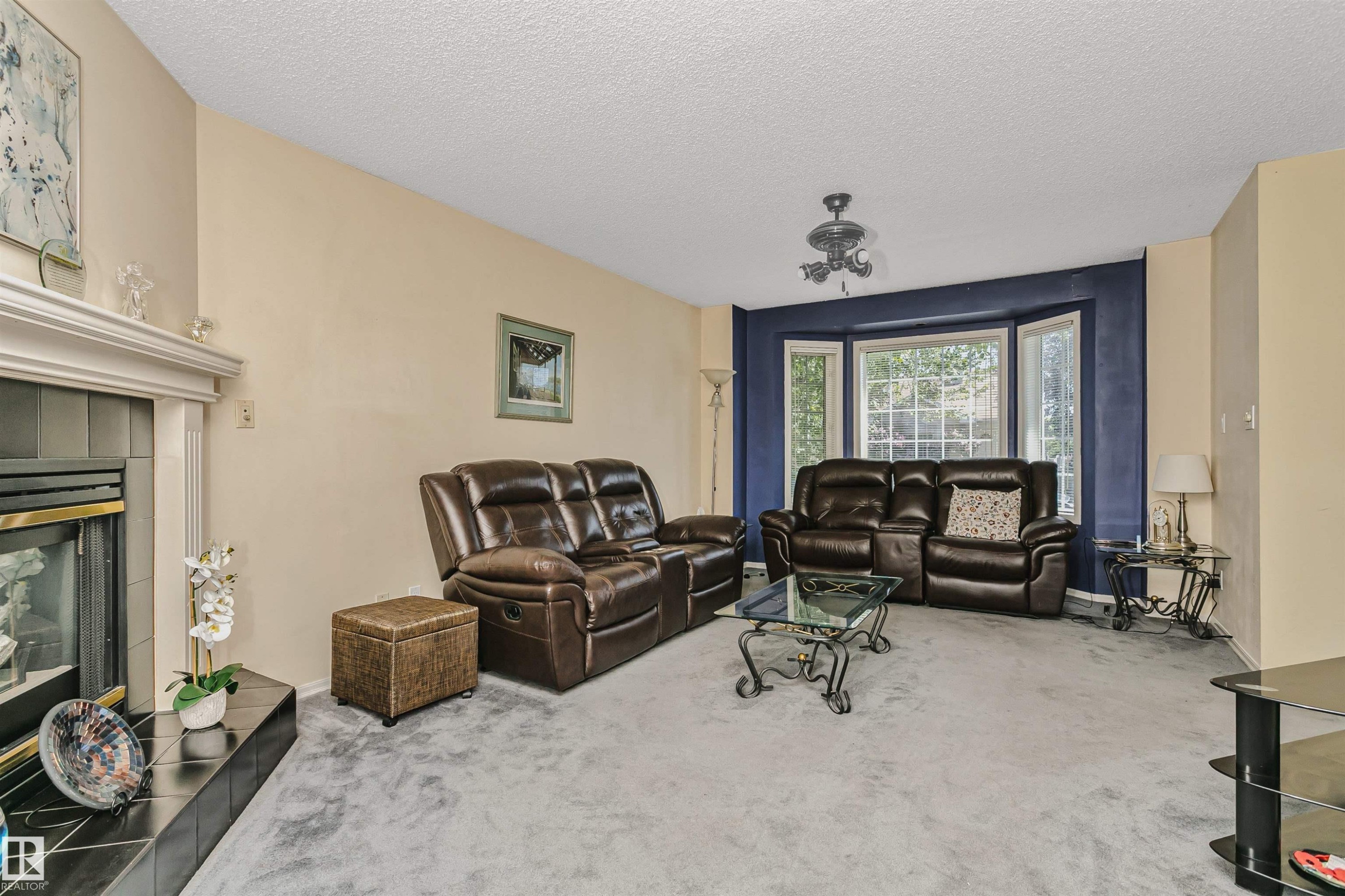 Living area featuring a fireplace with a black tile surround and white mantel, light-colored walls, bay window with a dark accent wall, and textured ceiling - 1612 80A Street, Edmonton, AB