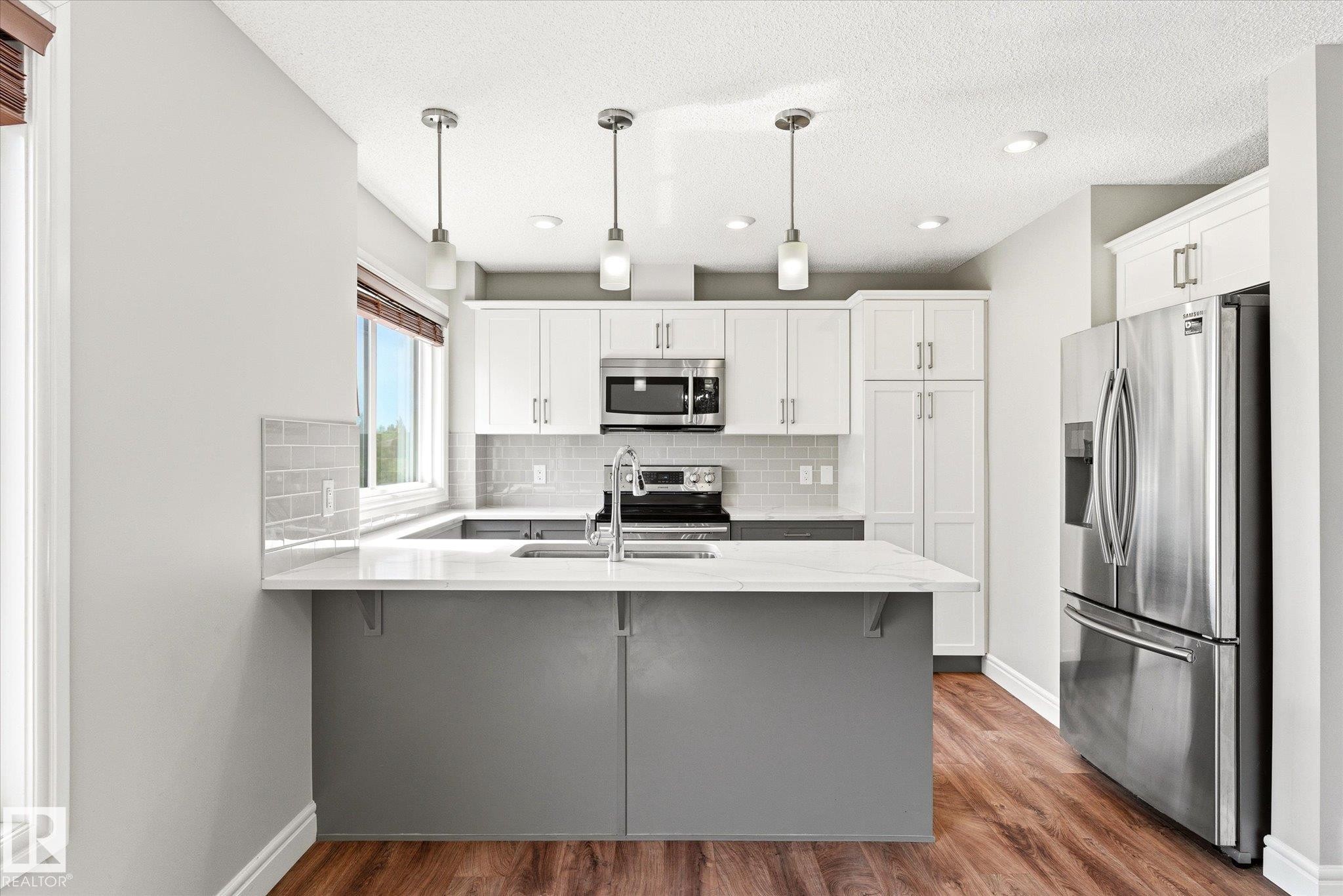 Modern kitchen featuring a central island with countertop seating, white shaker-style cabinetry, stainless steel appliances, a subway tile backsplash, and wood-finish flooring - 47 330 Bulyea Road, Edmonton, AB - Indoor Photo Showing Kitchen With Stainless Steel Kitchen With Upgraded Kitchen