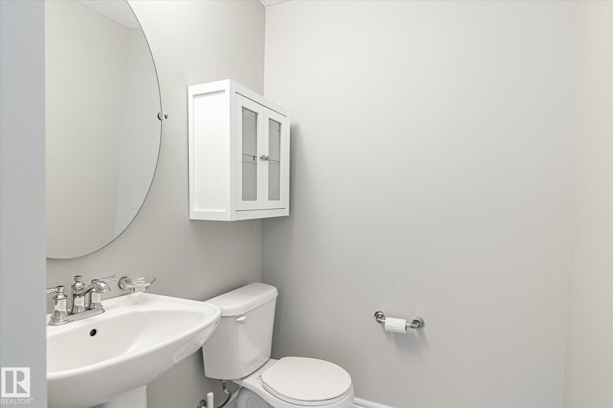 Pedestal sink with chrome faucet, circular wall mirror, and white wall cabinet with glass-paneled doors - 47 330 Bulyea Road, Edmonton, AB - Indoor Photo Showing Bathroom