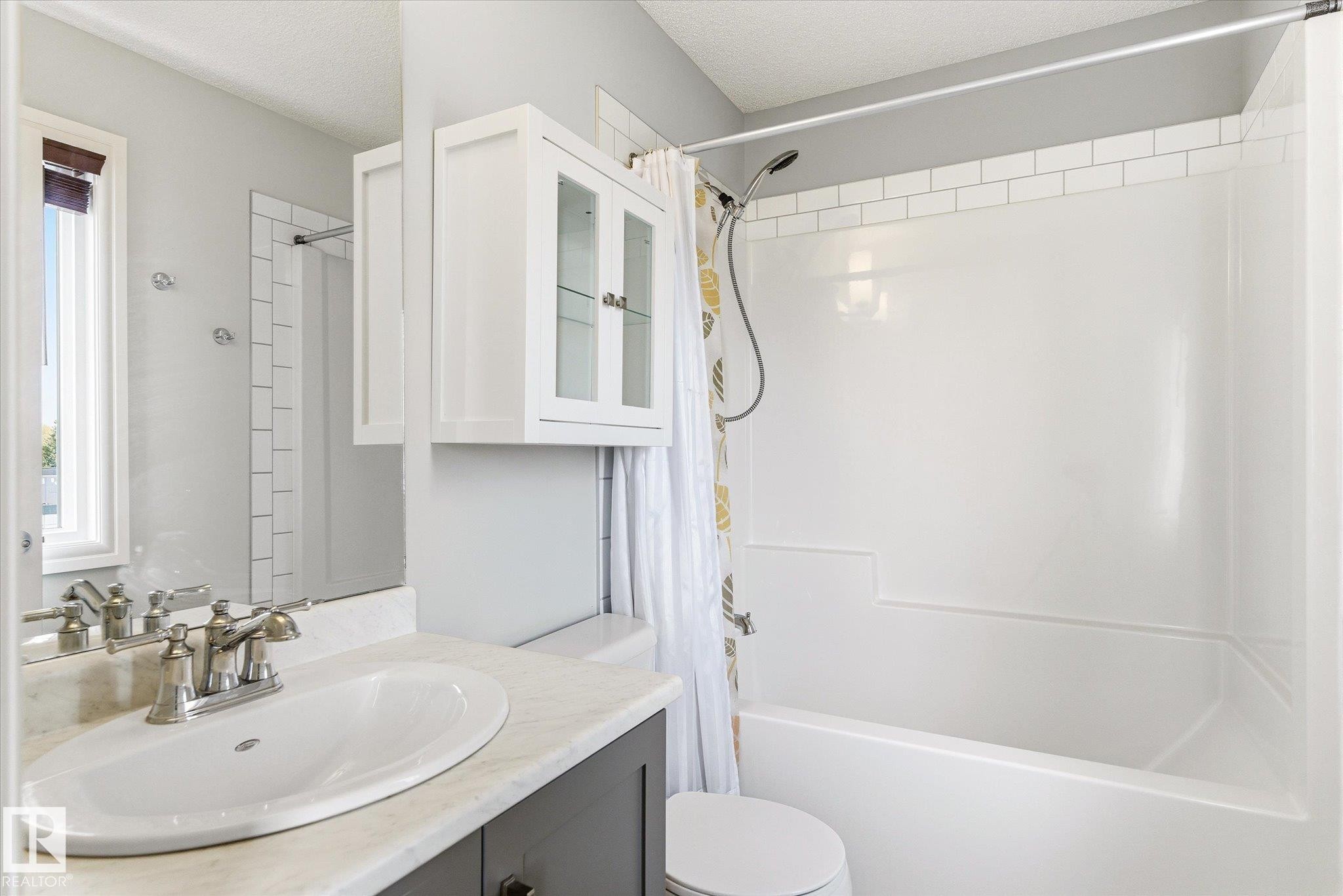 Bathroom featuring a single vanity with a white oval sink and chrome faucet, a wall-mounted medicine cabinet with glass-front doors, a window with a brown roll-down shade, and a bathtub with a white subway tile surround - 47 330 Bulyea Road, Edmonton, AB - Indoor Photo Showing Bathroom