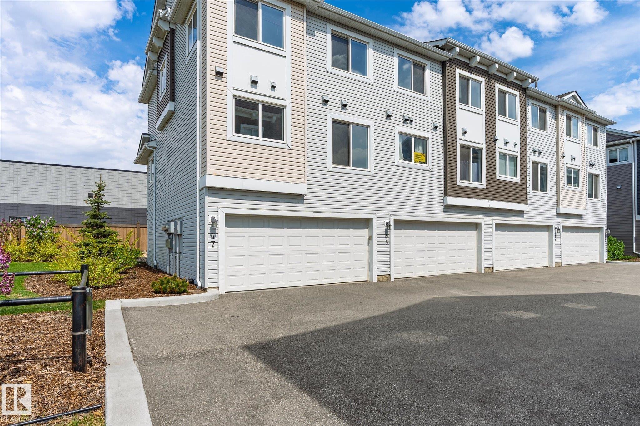 Multi-level townhouse exterior featuring light-colored siding, multiple windows, an attached garage with a white door, and a paved driveway - 47 330 Bulyea Road, Edmonton, AB - Outdoor