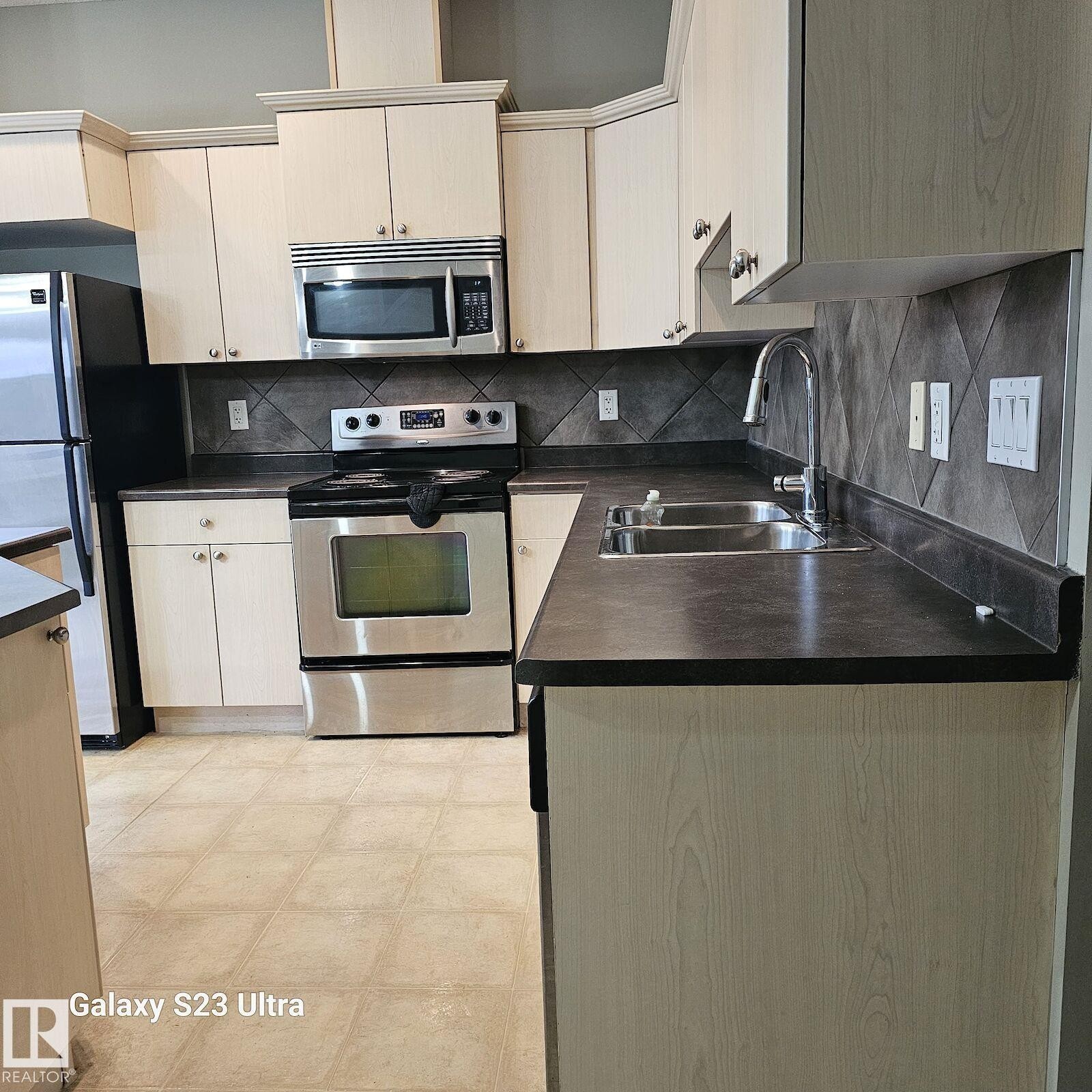 Kitchen featuring light wood-finish cabinetry, dark countertops, and a full suite of stainless steel appliances - 99 13825 155 Avenue, Edmonton, AB