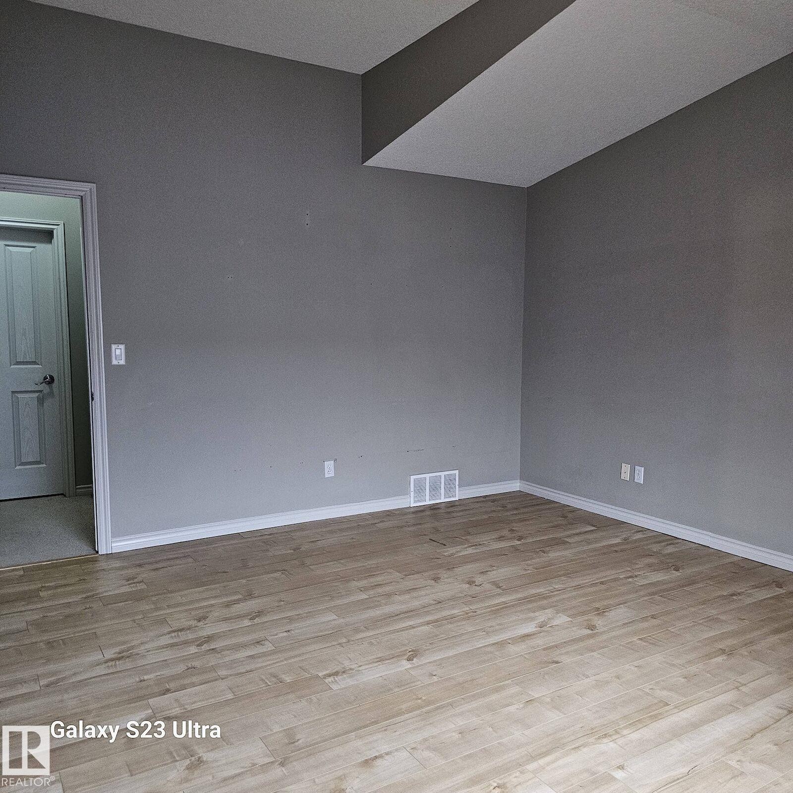 Spacious room featuring light wood-finish flooring, neutral gray wall paint, white trim, and a white interior door - 99 13825 155 Avenue, Edmonton, AB