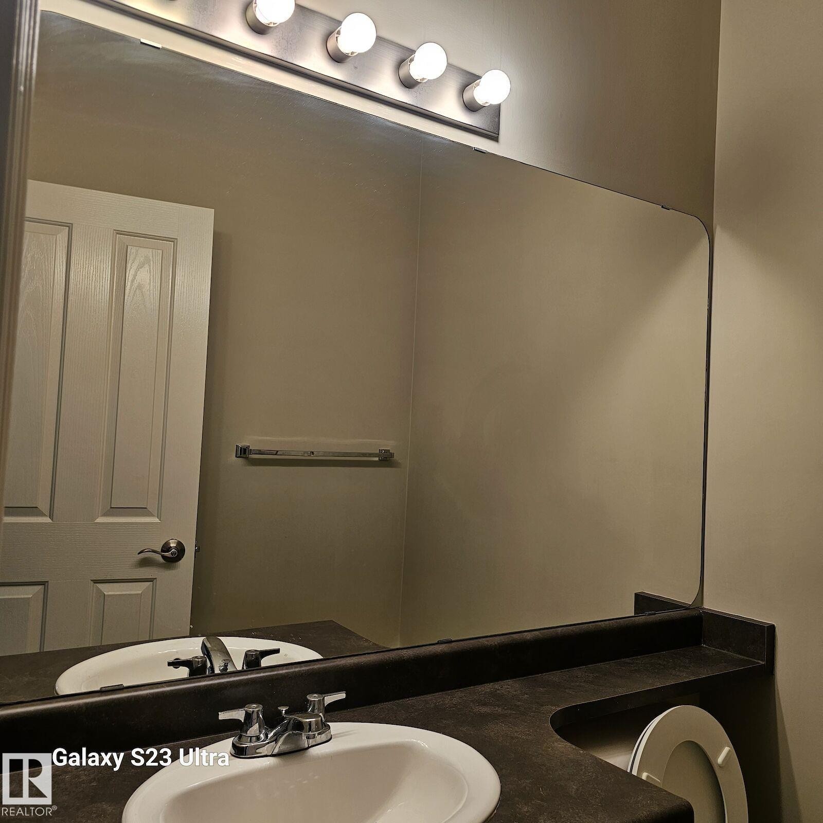 Bathroom vanity featuring a large wall-mounted mirror, brushed nickel light fixture with five bulbs, dark countertop with an integrated white oval sink, and a chrome two-handle faucet - 99 13825 155 Avenue, Edmonton, AB