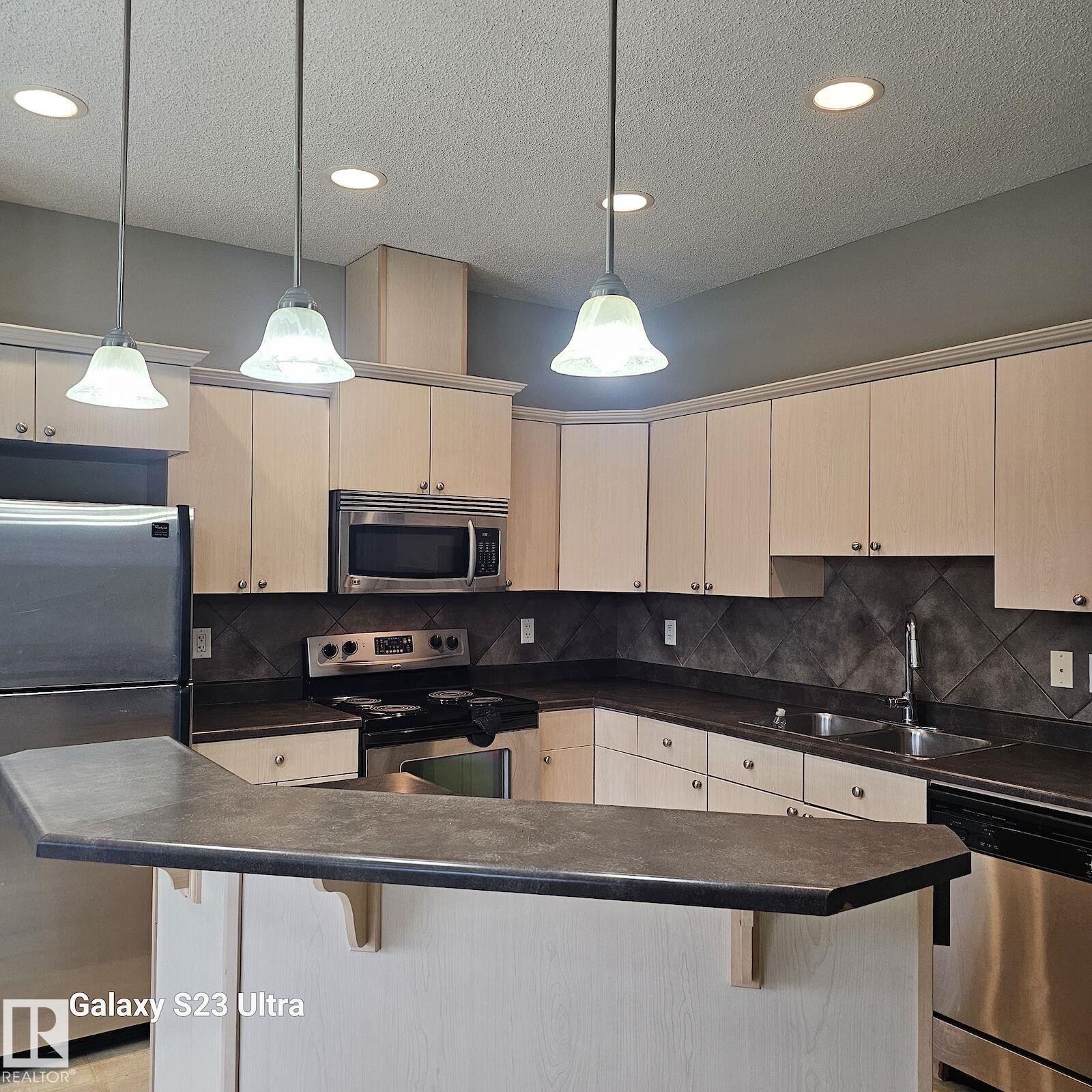 Kitchen featuring a peninsula breakfast bar, stainless steel appliances, light wood-finish cabinetry, dark countertops, and a tiled backsplash - 99 13825 155 Avenue, Edmonton, AB