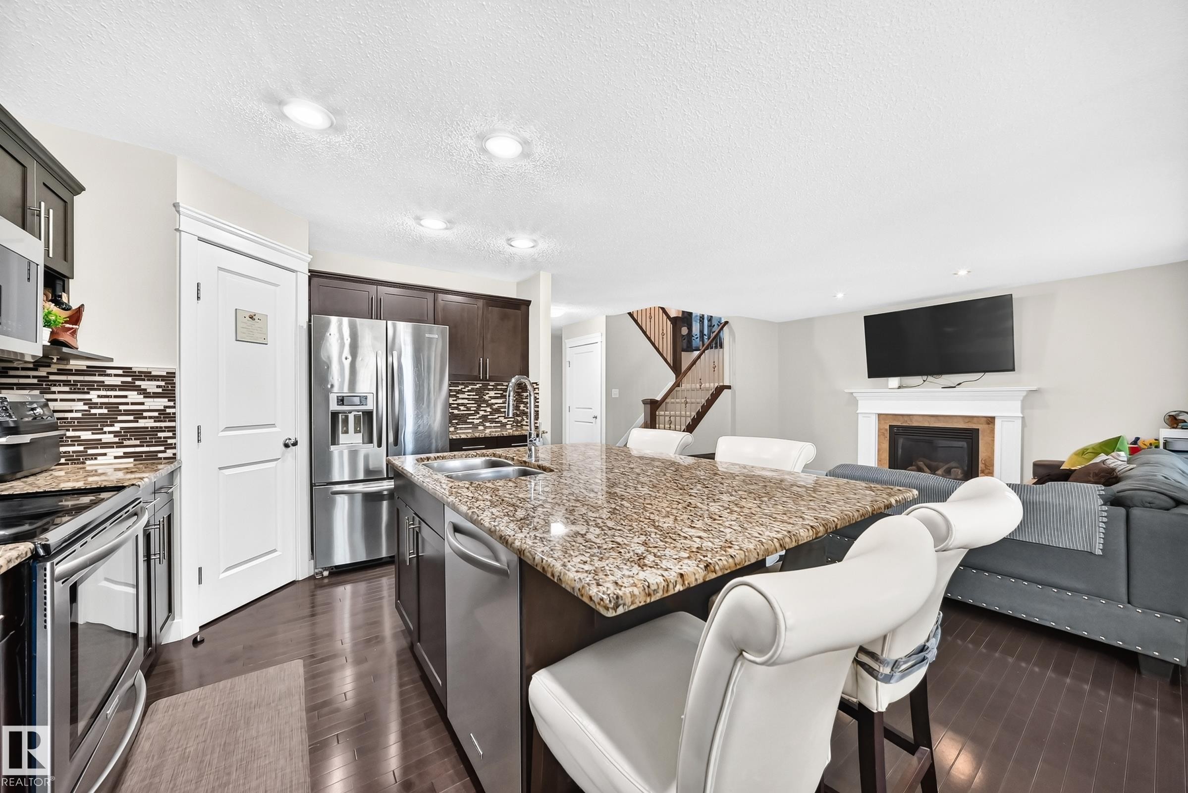 Open-concept kitchen featuring an island with granite countertop and double-basin sink, stainless steel appliances, dark wood cabinetry, and a mosaic tile backsplash - 3433 Goodridge Link Link, Edmonton, AB