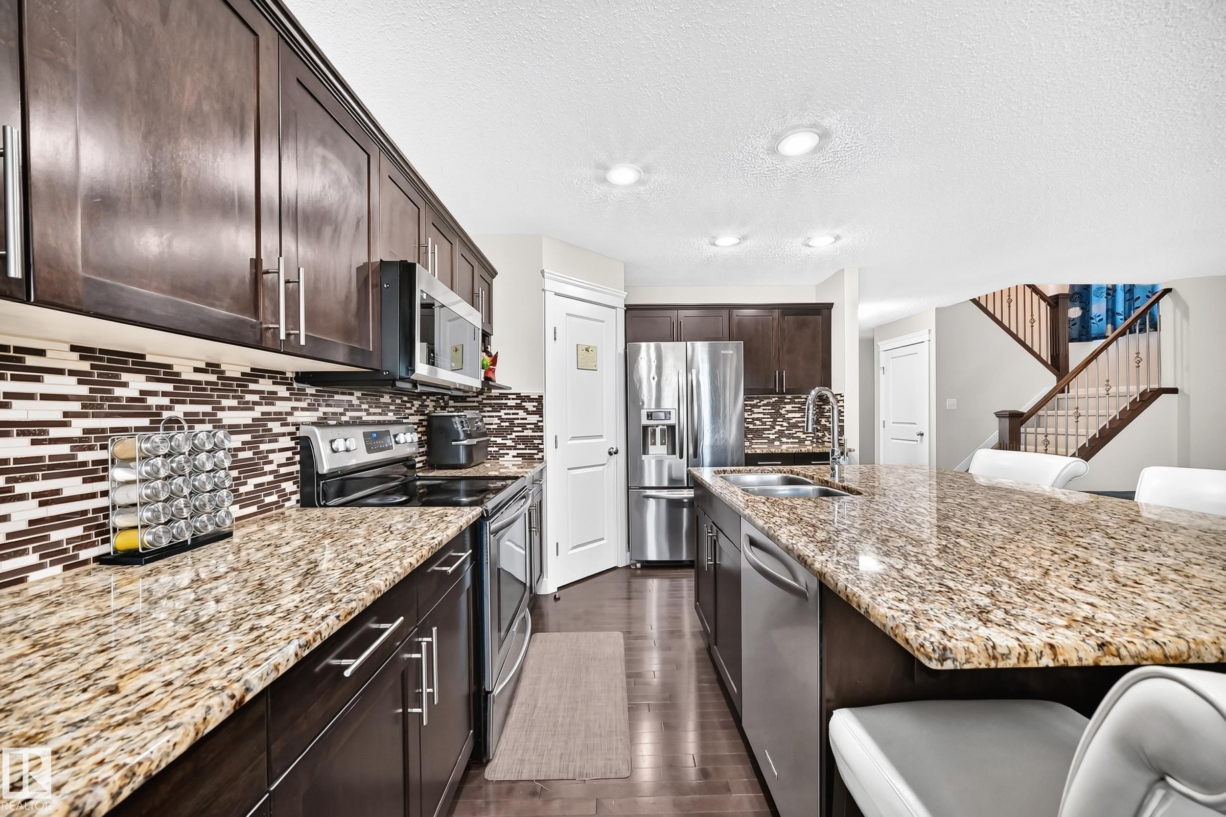 Well-appointed kitchen featuring dark wood cabinetry, stainless steel appliances, a mosaic tile backsplash, and extensive granite countertops including a large island with integrated sink - 3433 Goodridge Link Link, Edmonton, AB