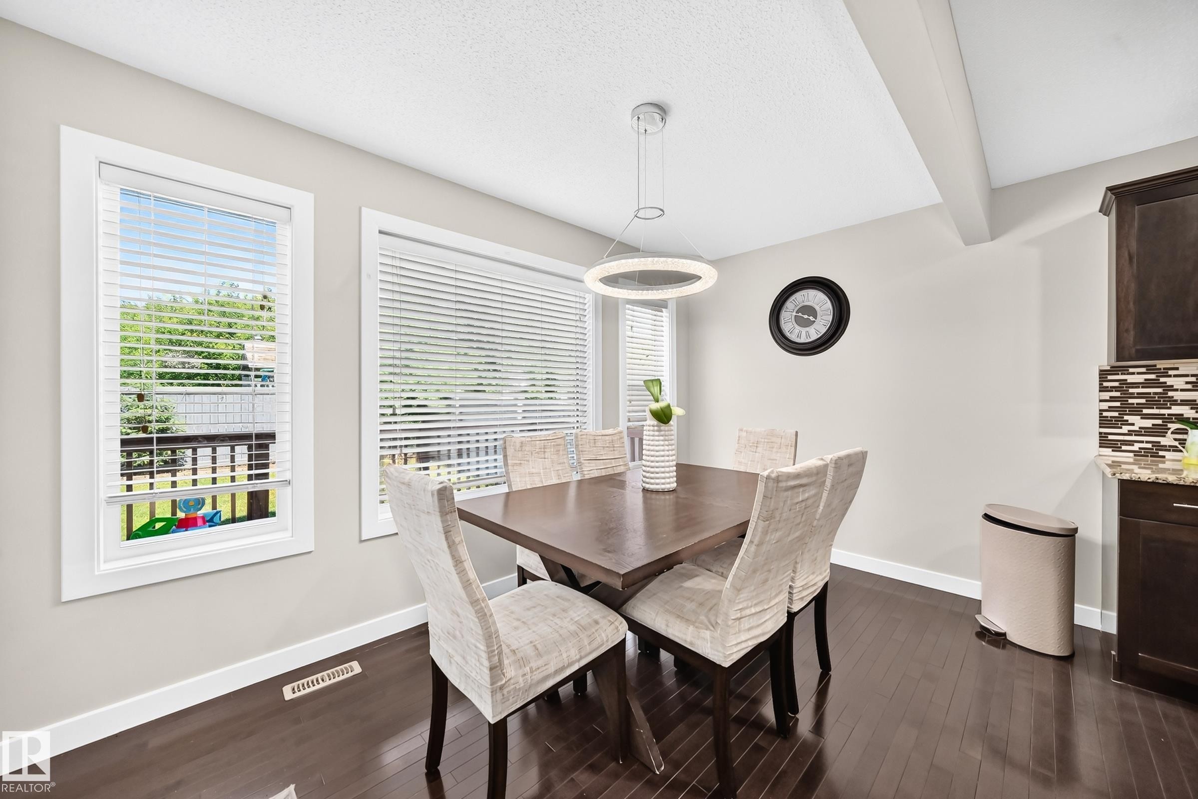 Dining area featuring wood-finish flooring, two large windows with blinds, a contemporary pendant light fixture, and a partial view of kitchen cabinetry with a mosaic tile backsplash - 3433 Goodridge Link Link, Edmonton, AB
