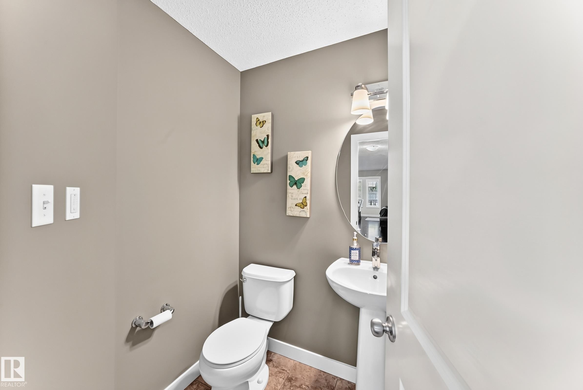 Compact powder room featuring a white pedestal sink, circular wall mirror, and two-light vanity fixture - 3433 Goodridge Link Link, Edmonton, AB