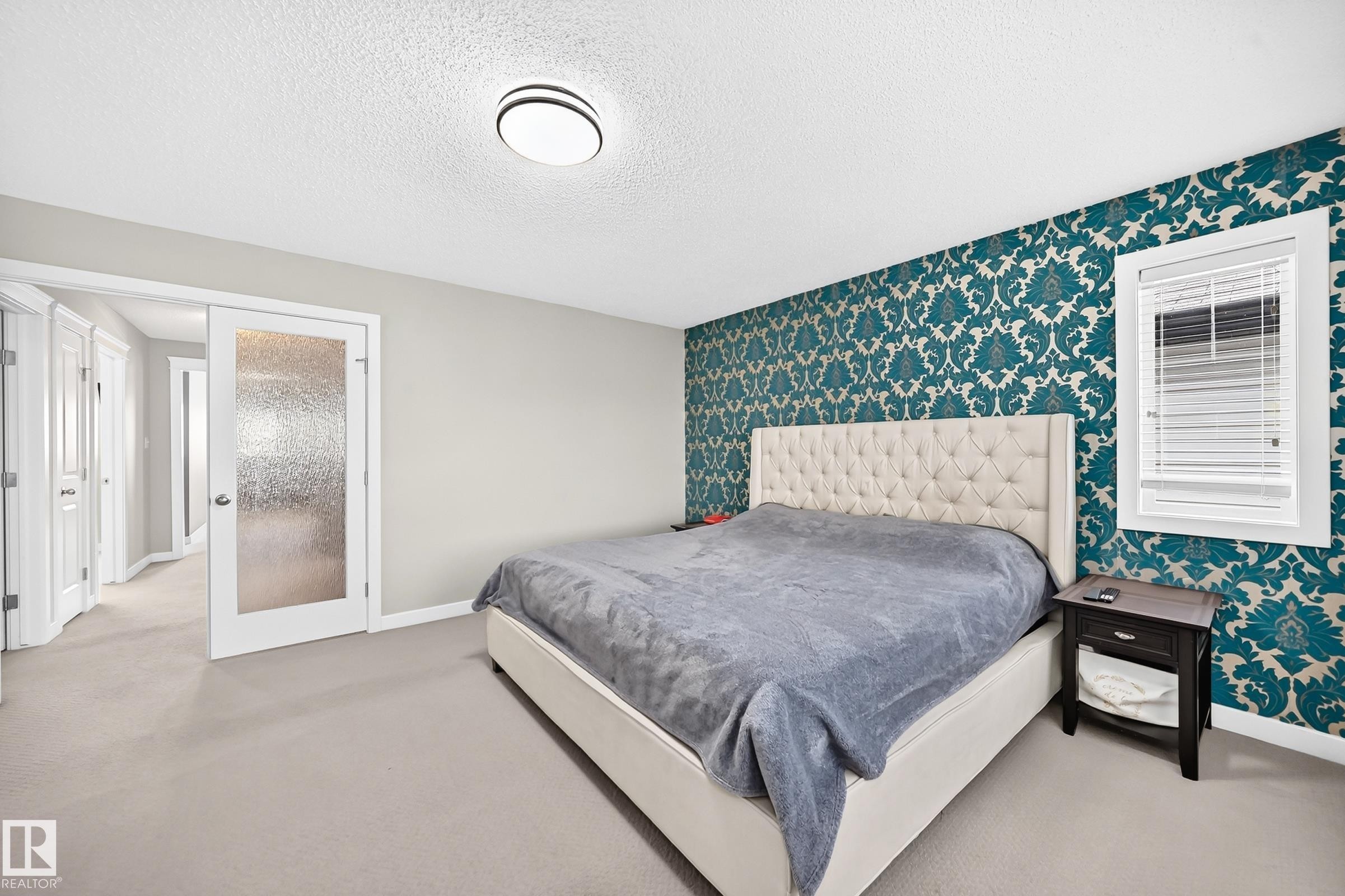 Bedroom featuring an accent wall with teal and gold patterned wallpaper, light gray carpeting, and a white window frame with horizontal blinds - 3433 Goodridge Link Link, Edmonton, AB