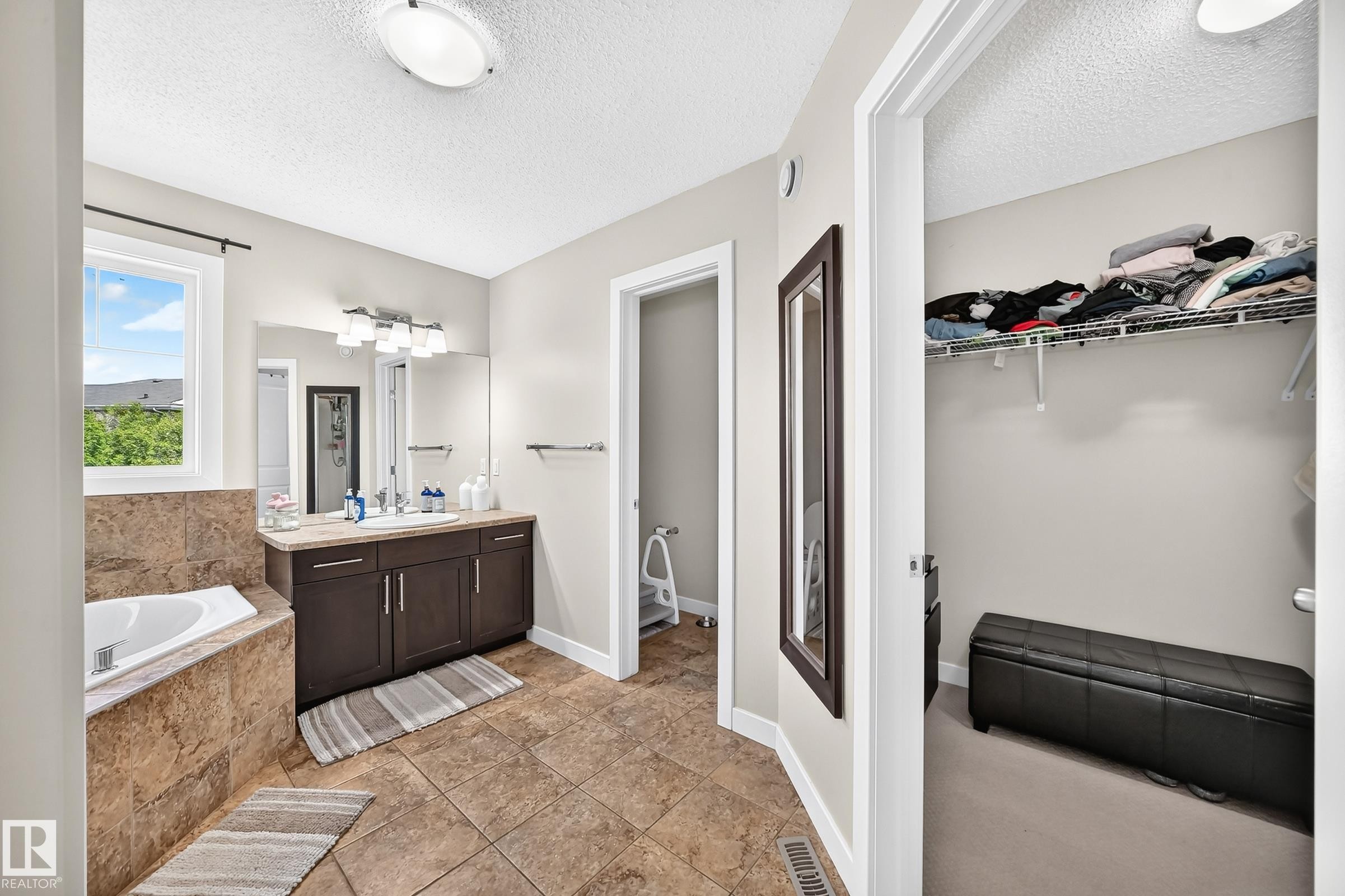 Bathroom featuring a built-in soaking tub with a tiled surround, a dark wood-finish vanity with a light-colored countertop and double sinks, and ceramic tile flooring - 3433 Goodridge Link Link, Edmonton, AB