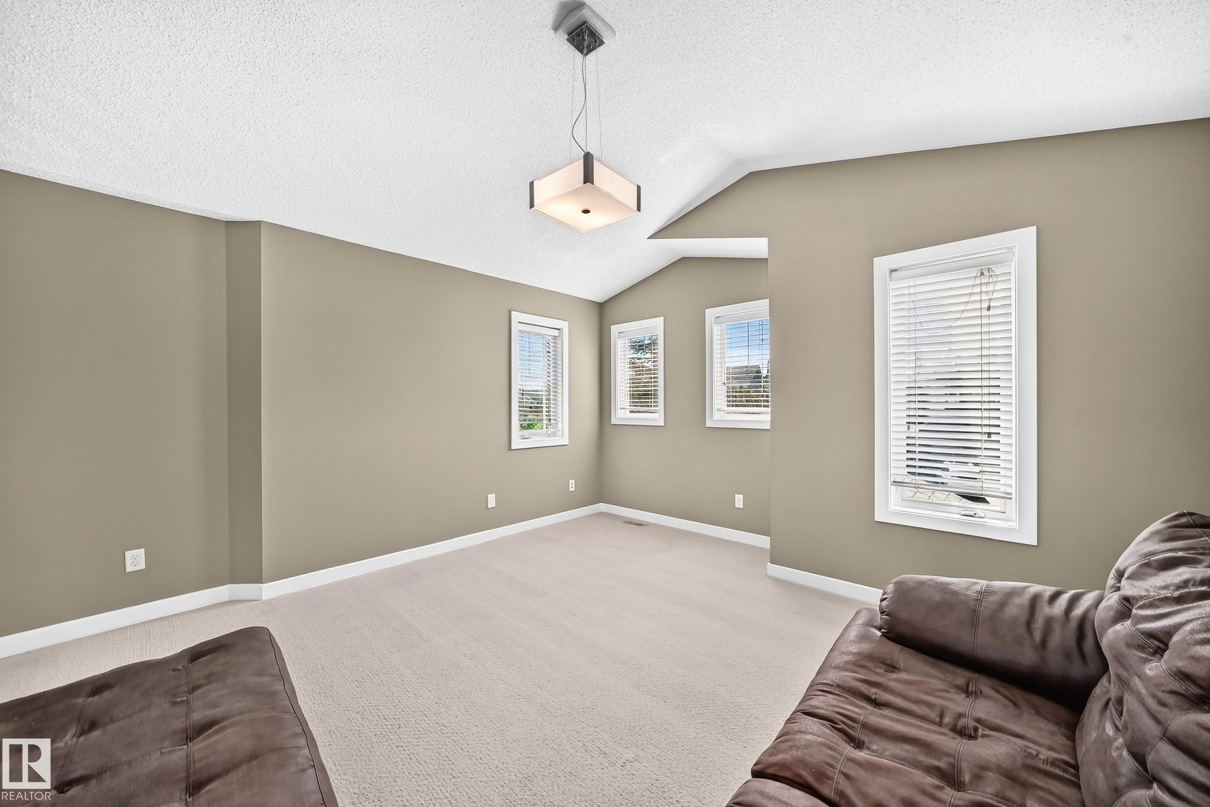 Spacious room featuring vaulted ceilings, a contemporary ceiling light fixture, multiple windows with white blinds, neutral wall tones, and light-colored carpeting - 3433 Goodridge Link Link, Edmonton, AB