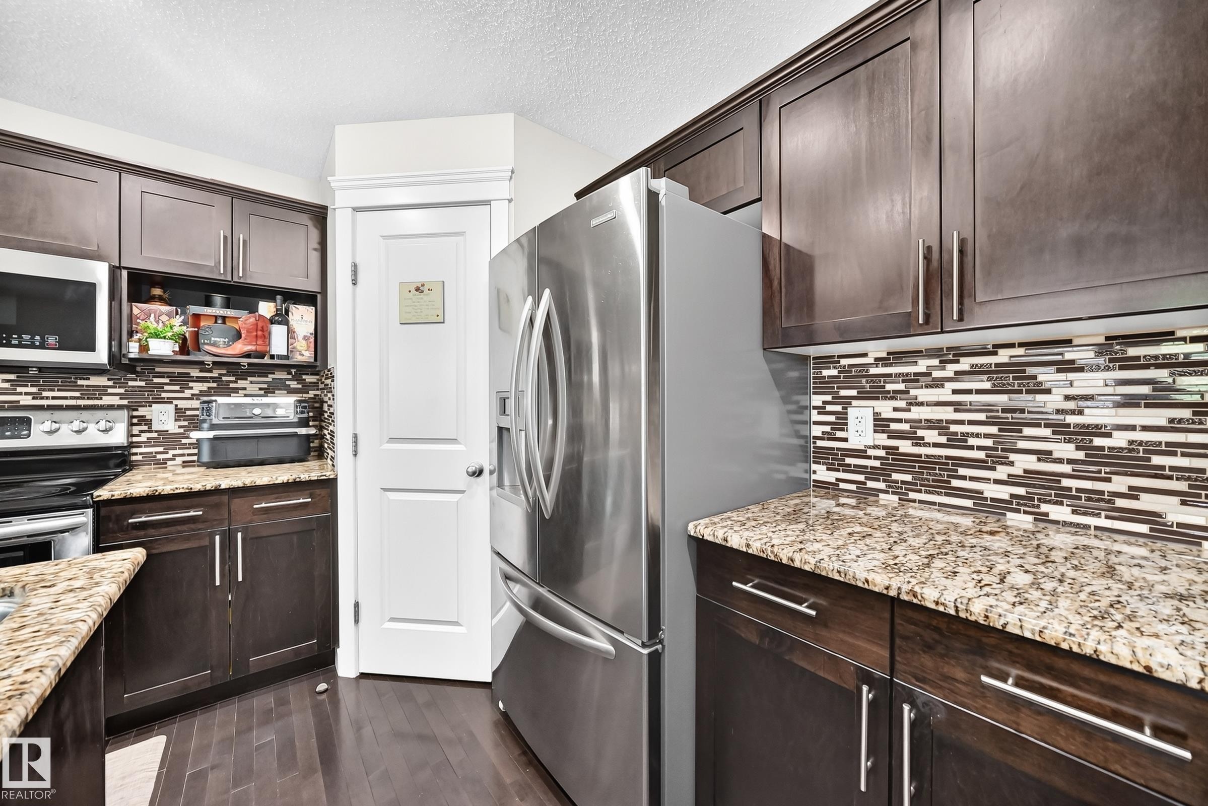 Kitchen featuring rich wood-finish cabinetry, speckled countertops, a mosaic tile backsplash, and stainless steel appliances including a refrigerator and built-in microwave - 3433 Goodridge Link Link, Edmonton, AB