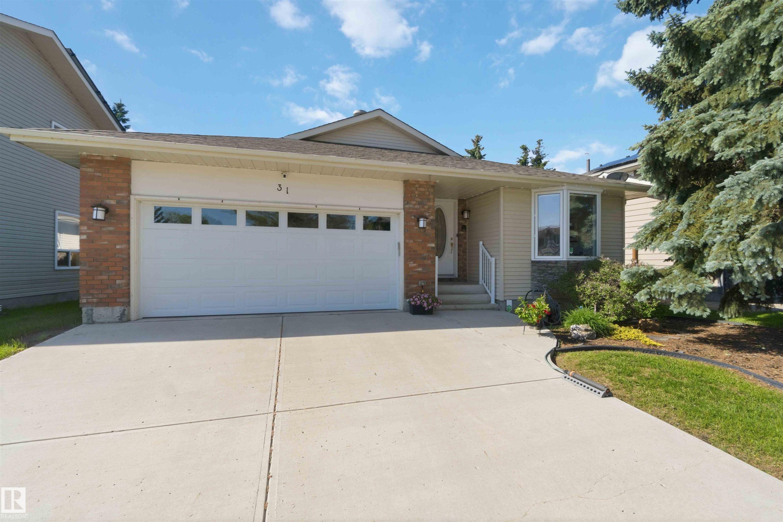 Exterior featuring a two-car garage with transom windows, a concrete driveway, mixed material facade with brick and stone accents, a covered front entry with white railings, and a prominent front window - 31 Gariepy Crescent, Edmonton, AB