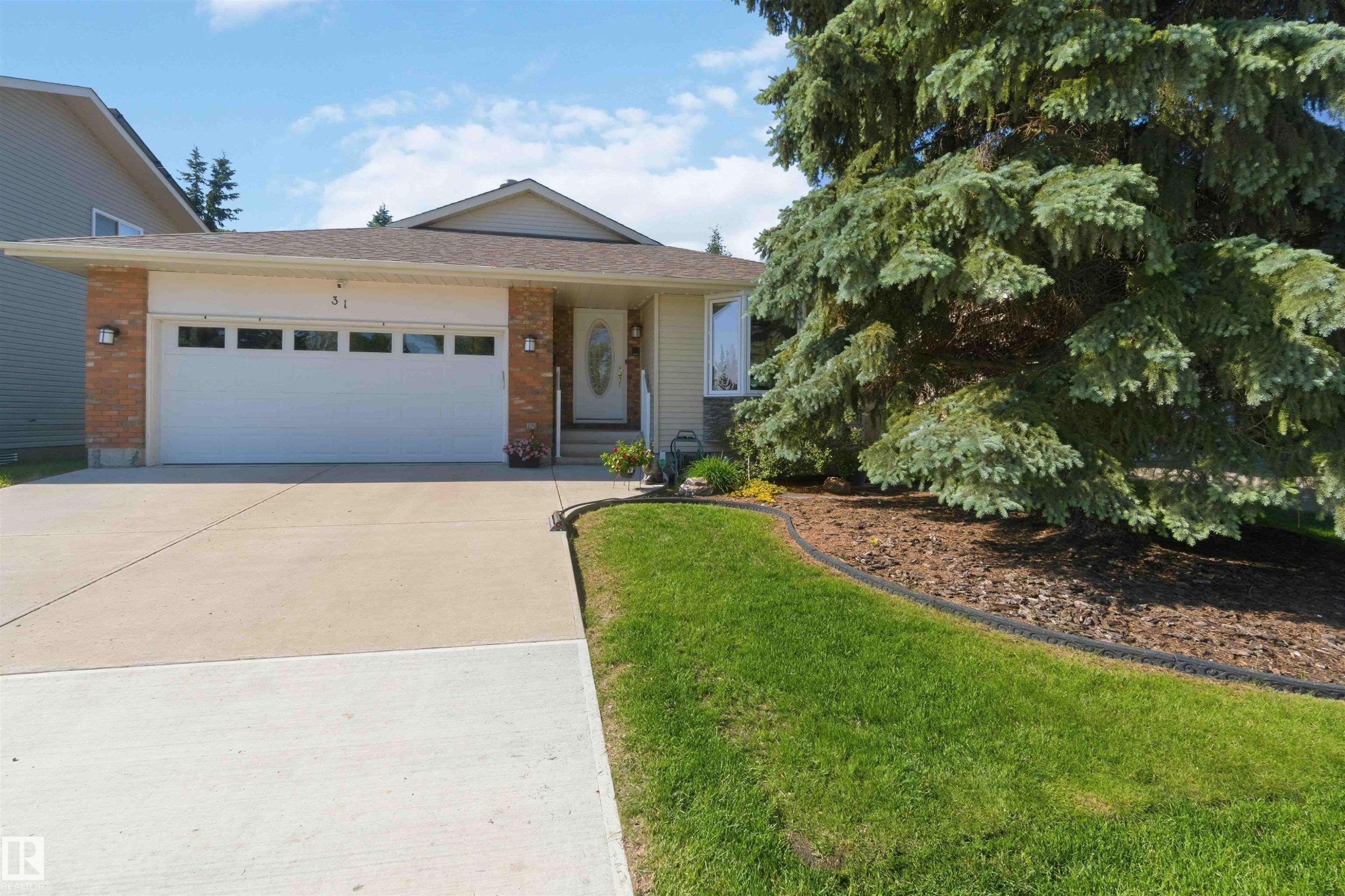 Exterior featuring a two-car garage with frosted window panels, brick accents, and a mature evergreen tree - 31 Gariepy Crescent, Edmonton, AB