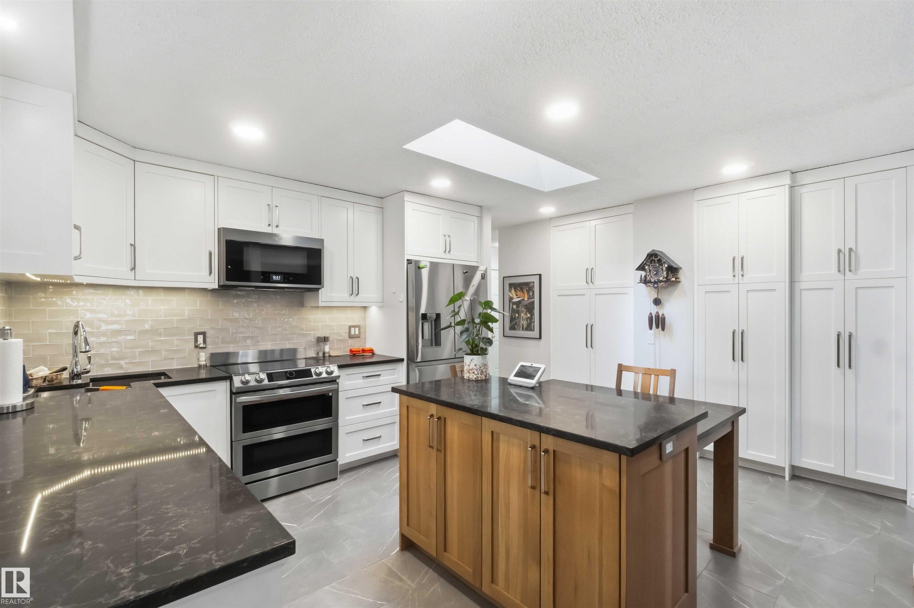 Bright kitchen featuring white cabinetry, dark countertops, stainless steel appliances, a wood-finish island, and a skylight - 31 Gariepy Crescent, Edmonton, AB
