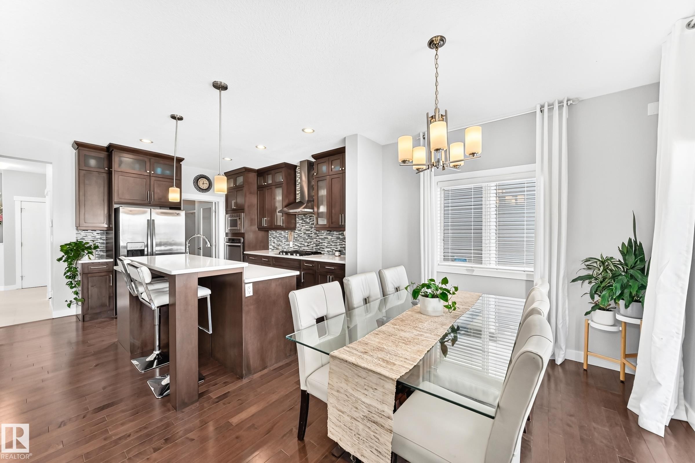 Open-concept living space featuring wood-finish flooring, a kitchen island with seating, dark wood cabinetry, stainless steel appliances, and a dining area with a window - 3672 Claxton Place Sw, Edmonton, AB