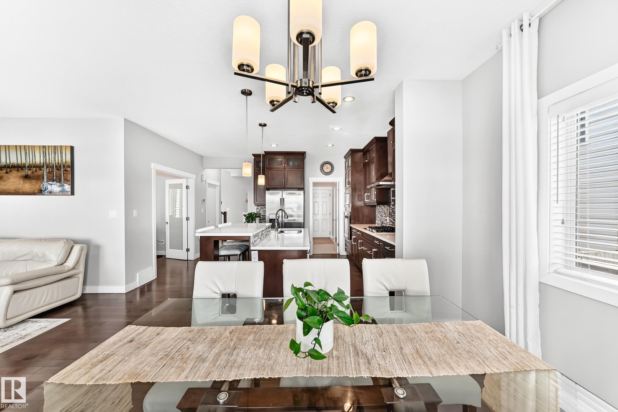 Open-concept interior featuring wood-finish flooring, a contemporary chandelier, a kitchen with dark wood cabinetry and light countertops, a kitchen island with pendant lighting, and a window with white blinds - 3672 Claxton Place Sw, Edmonton, AB