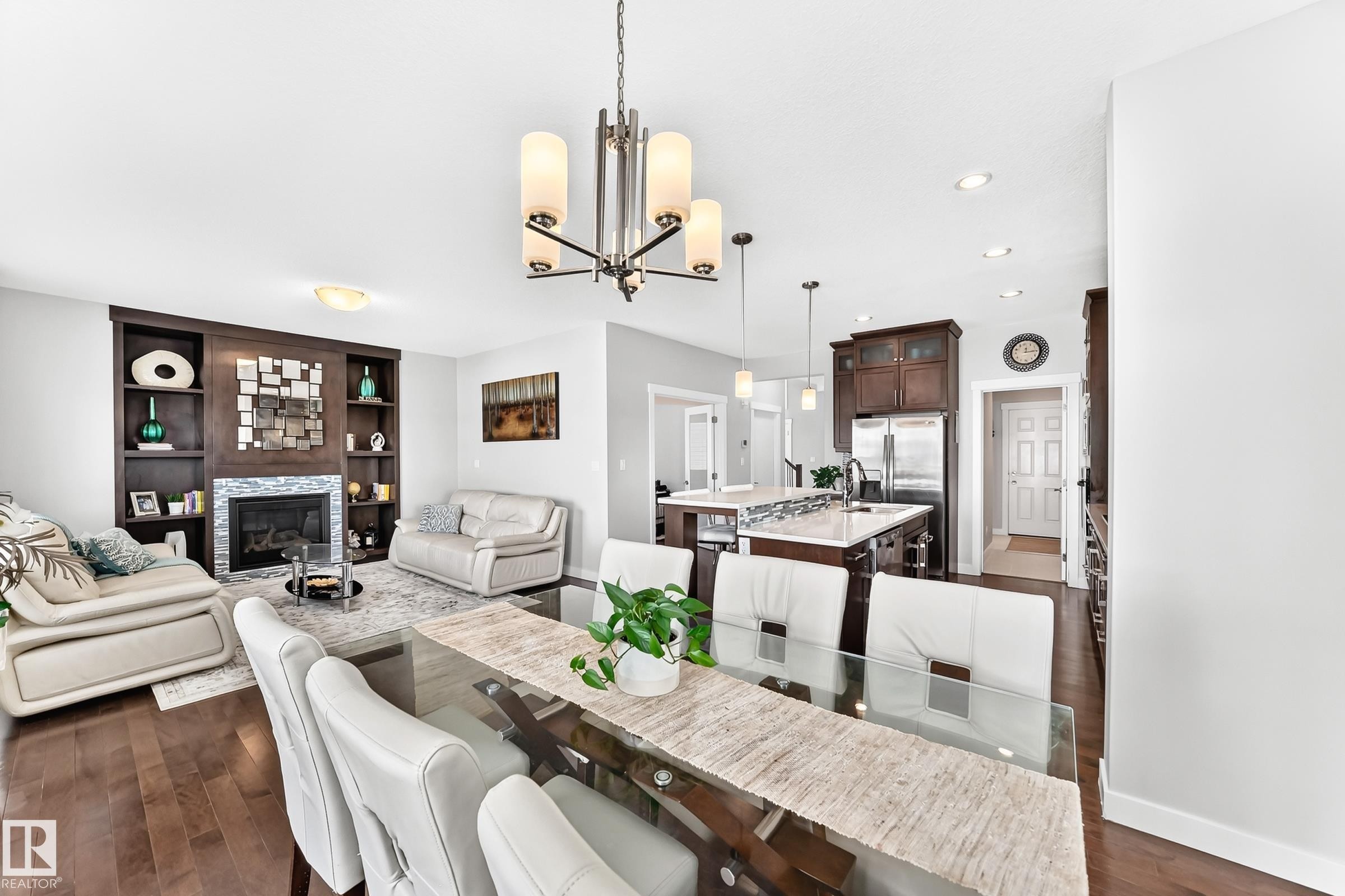 Open-concept living area featuring a central dining space, fireplace with stone surround and built-in shelving, hardwood-finish flooring, recessed lighting, and a kitchen with dark wood cabinetry and a central island - 3672 Claxton Place Sw, Edmonton, AB