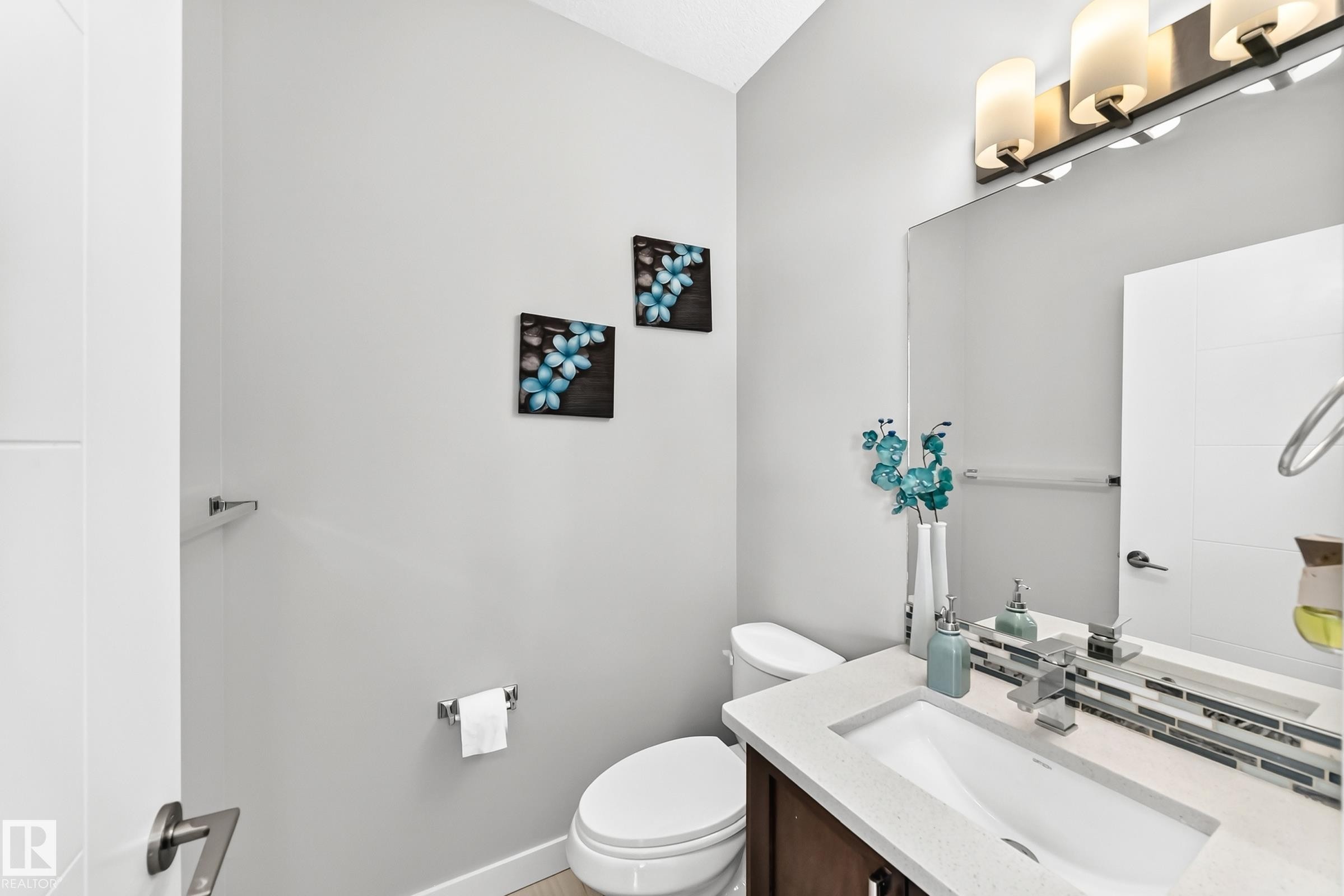 Powder room featuring a single vanity with a white countertop and integrated sink, a rectangular mirror, a three-light vanity fixture, and a tile backsplash - 3672 Claxton Place Sw, Edmonton, AB