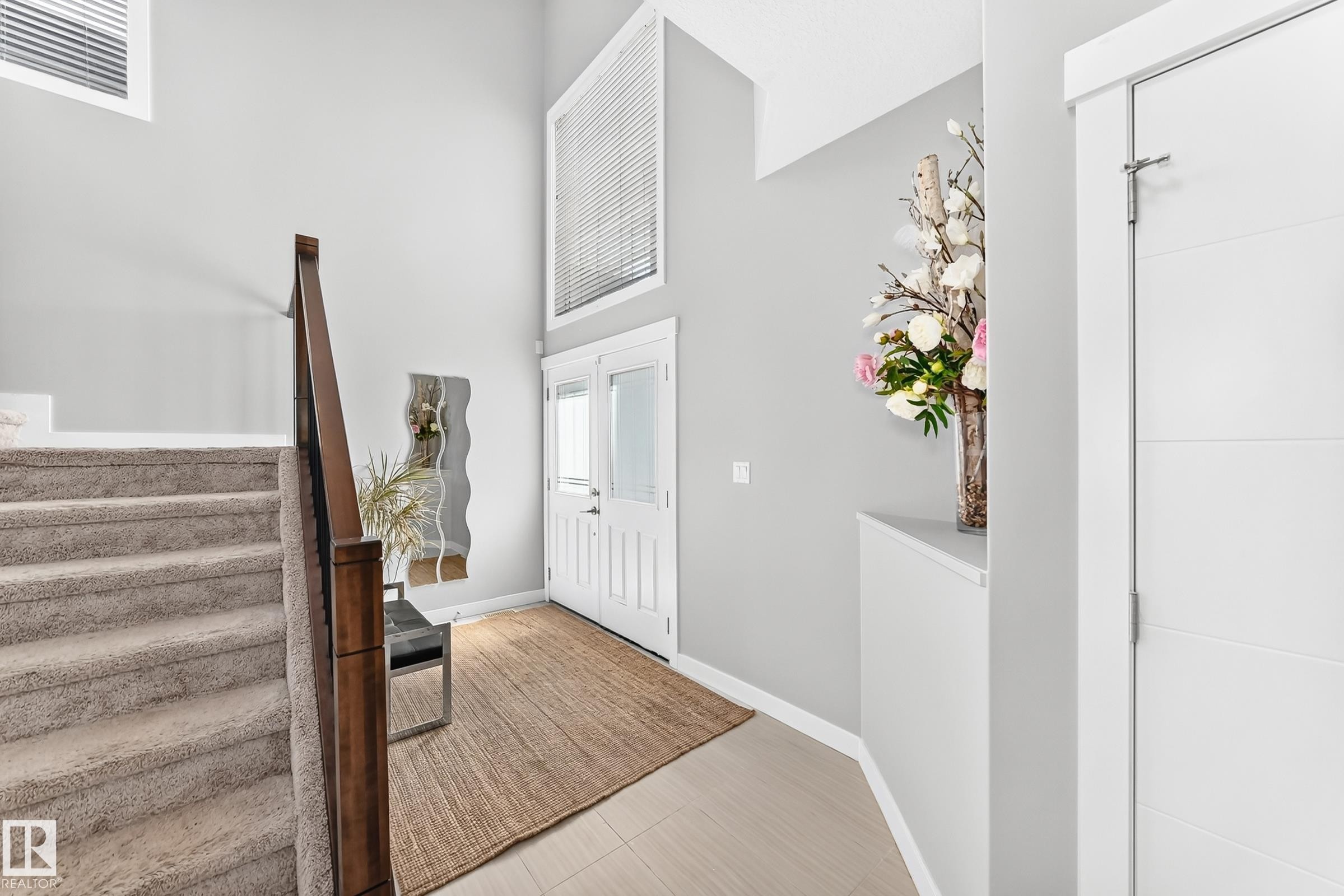 Two-story entryway featuring a carpeted staircase with wood banister, double front doors with glass panels, light wood-finish flooring, and a window with blinds positioned at a height - 3672 Claxton Place Sw, Edmonton, AB
