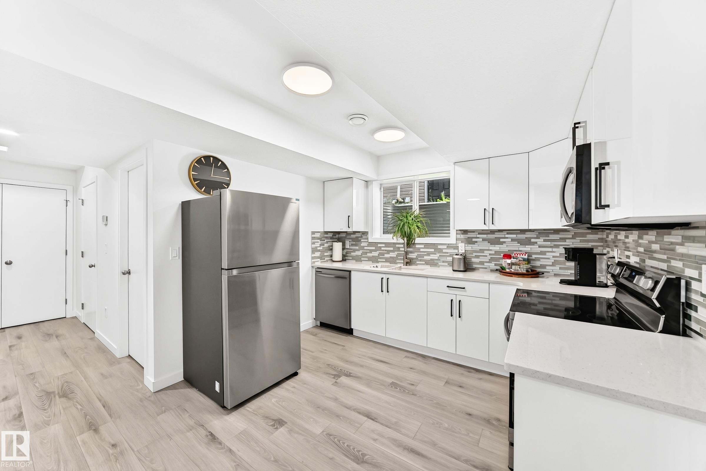 Modern kitchen featuring white cabinetry, light wood-finish flooring, stainless steel appliances, and a mosaic tile backsplash - 3672 Claxton Place Sw, Edmonton, AB