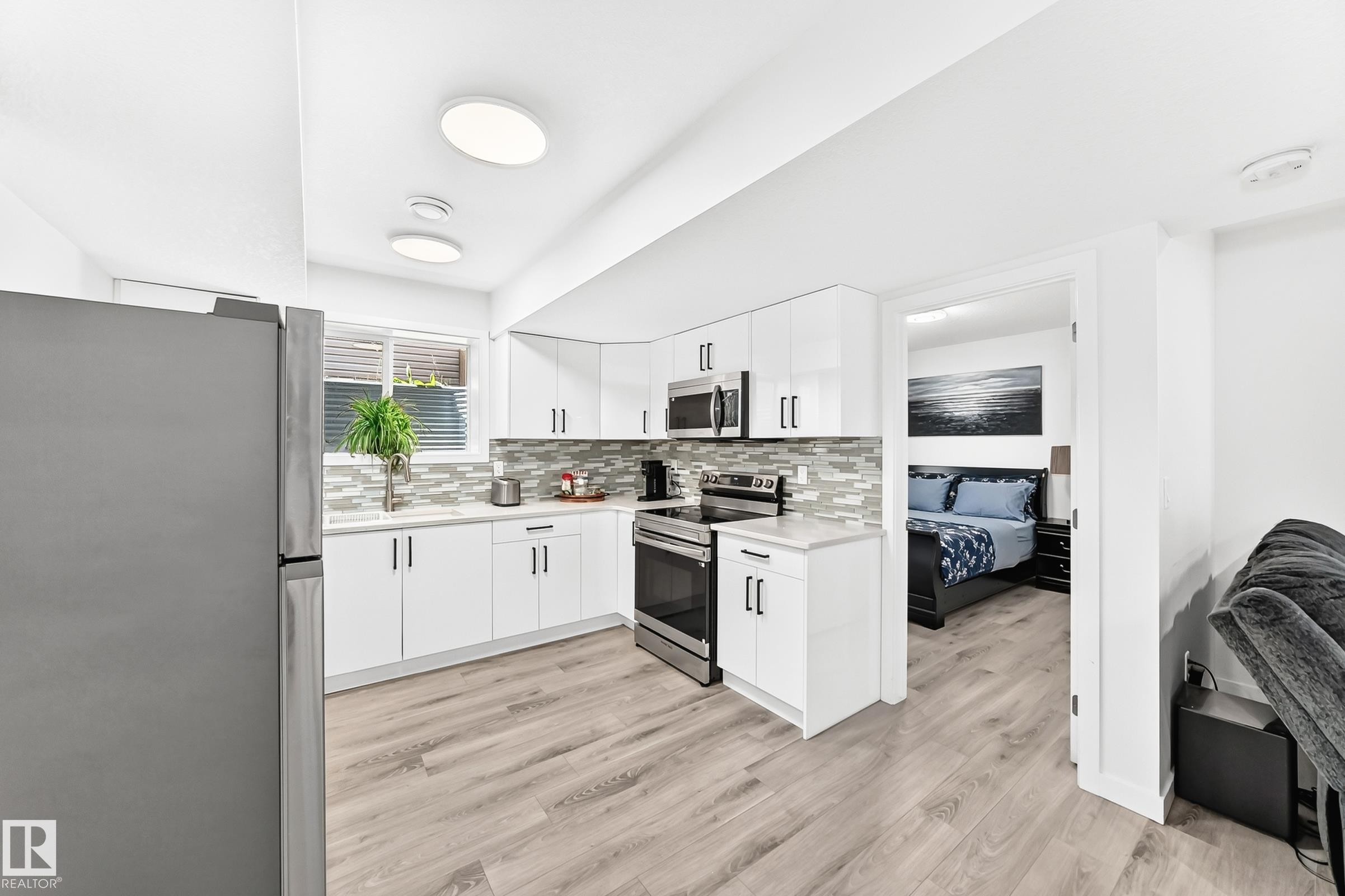 Modern kitchen featuring white cabinetry, light-toned countertops, a mosaic tile backsplash, stainless steel appliances, and wood-finish flooring - 3672 Claxton Place Sw, Edmonton, AB
