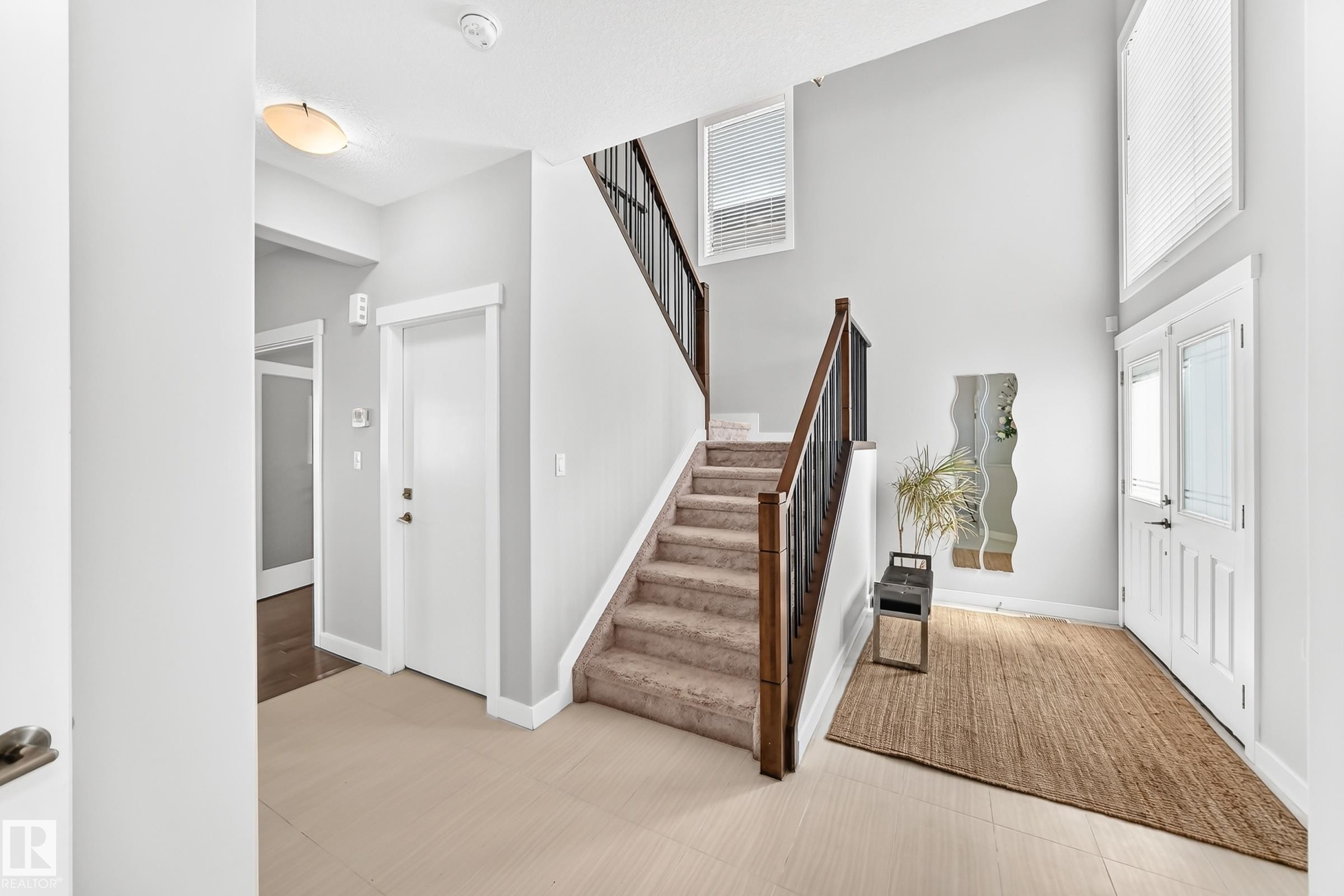 Spacious entry foyer featuring wood-finish flooring, a carpeted staircase with dark wood handrails and black balusters, and a double-door entryway with upper window panels - 3672 Claxton Place Sw, Edmonton, AB