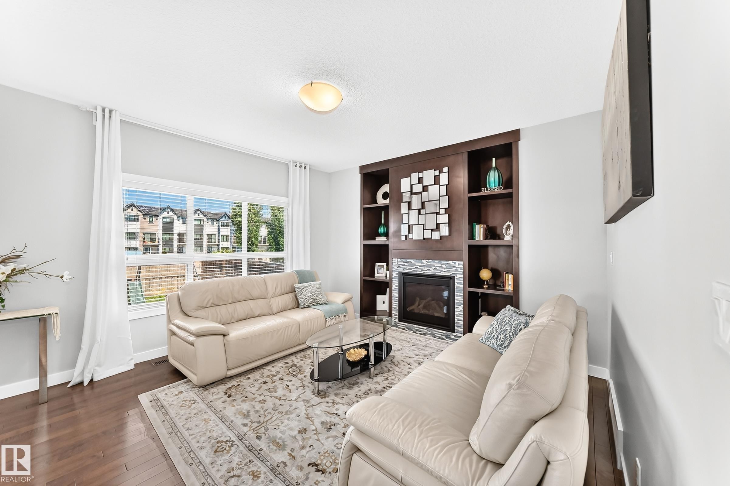 Living area featuring a built-in dark wood entertainment unit with shelving, a recessed fireplace with tile surround, and large windows with privacy blinds - 3672 Claxton Place Sw, Edmonton, AB