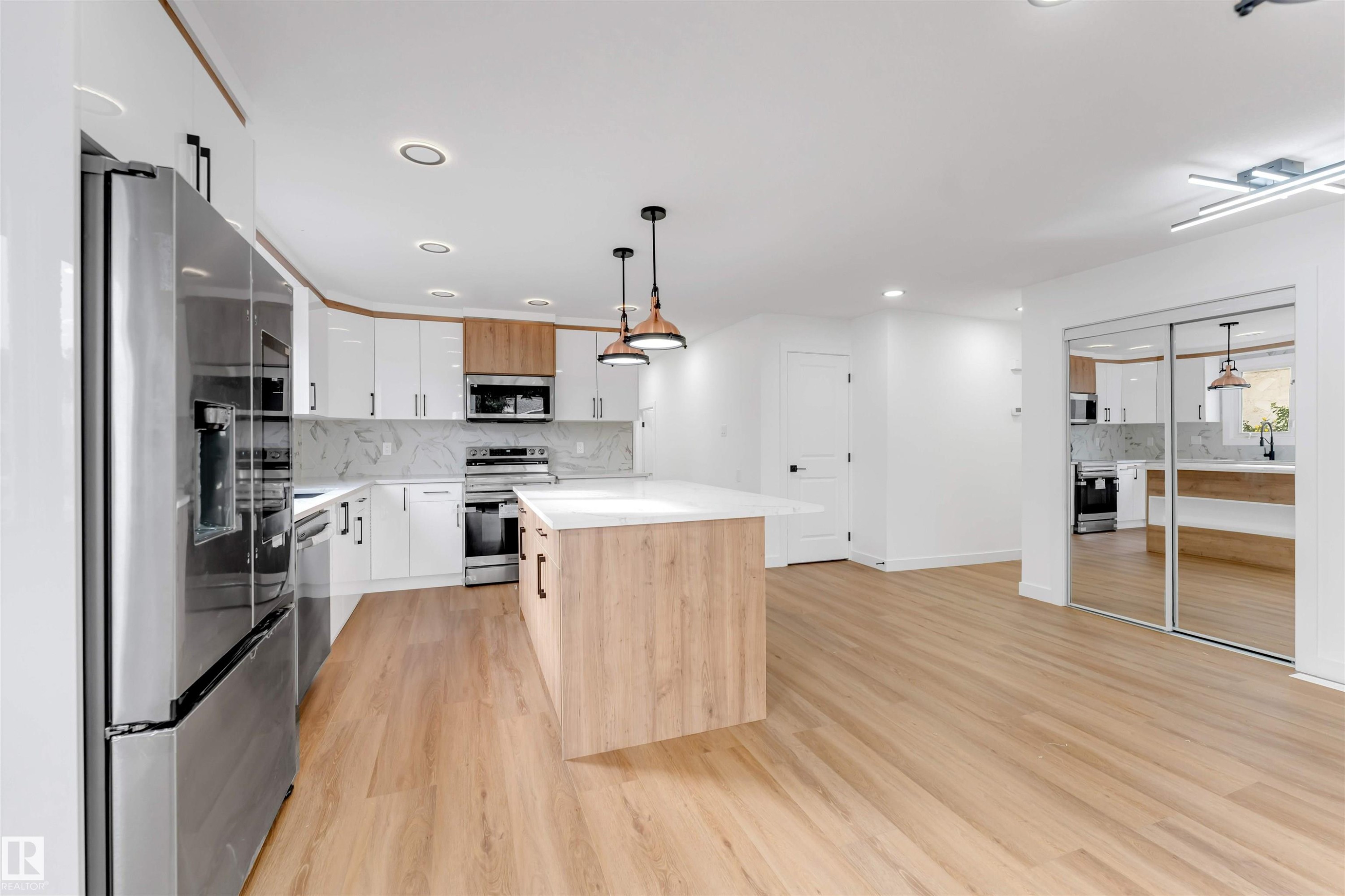 Contemporary kitchen featuring a white countertop island with wood-finish paneling, stainless steel appliances, white cabinetry with black pulls, and wood-finish flooring - 5512 150 Avenue, Edmonton, AB