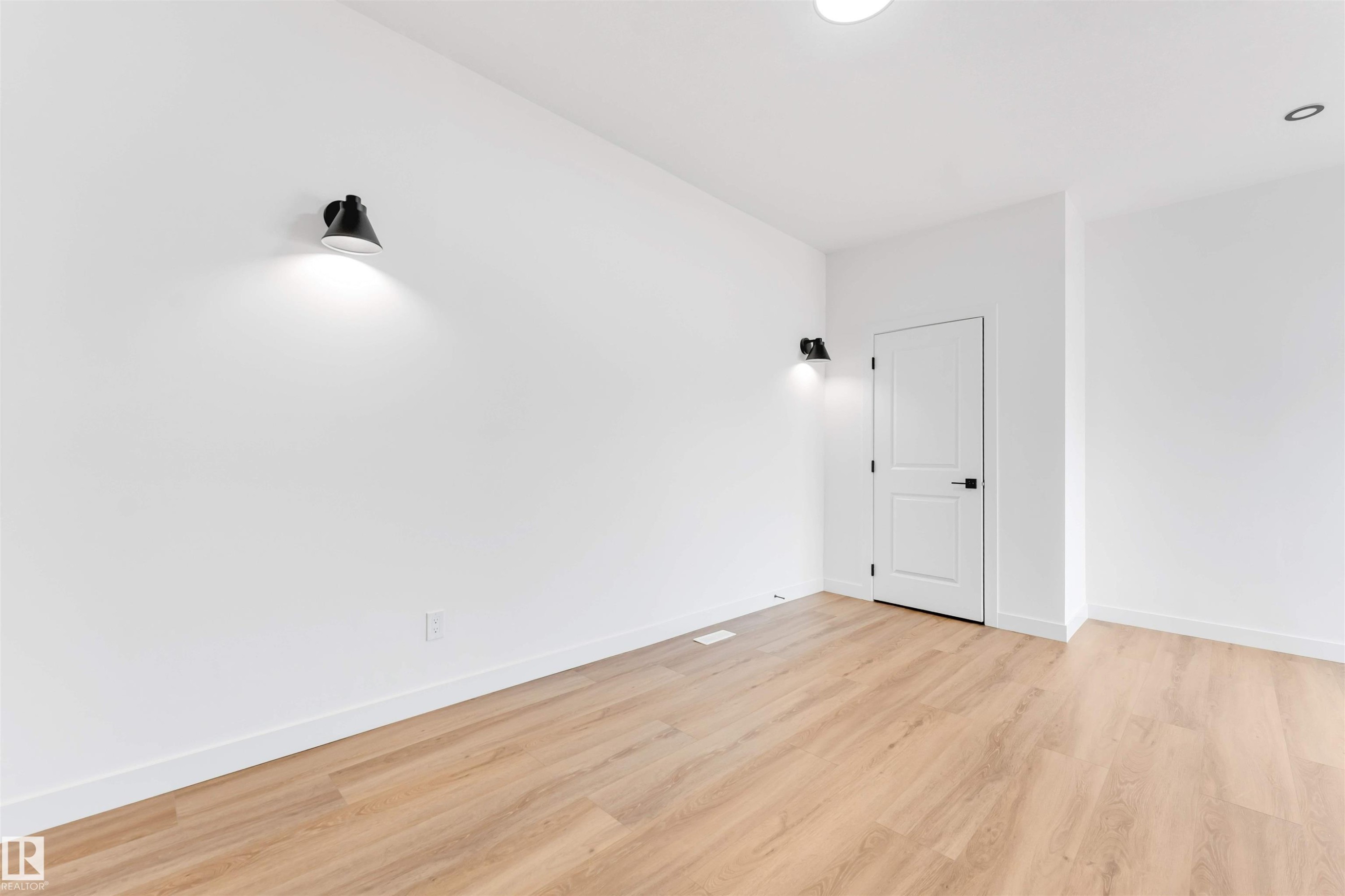 Spacious room featuring light wood-finish flooring, white walls, and contemporary black sconce lighting - 5512 150 Avenue, Edmonton, AB