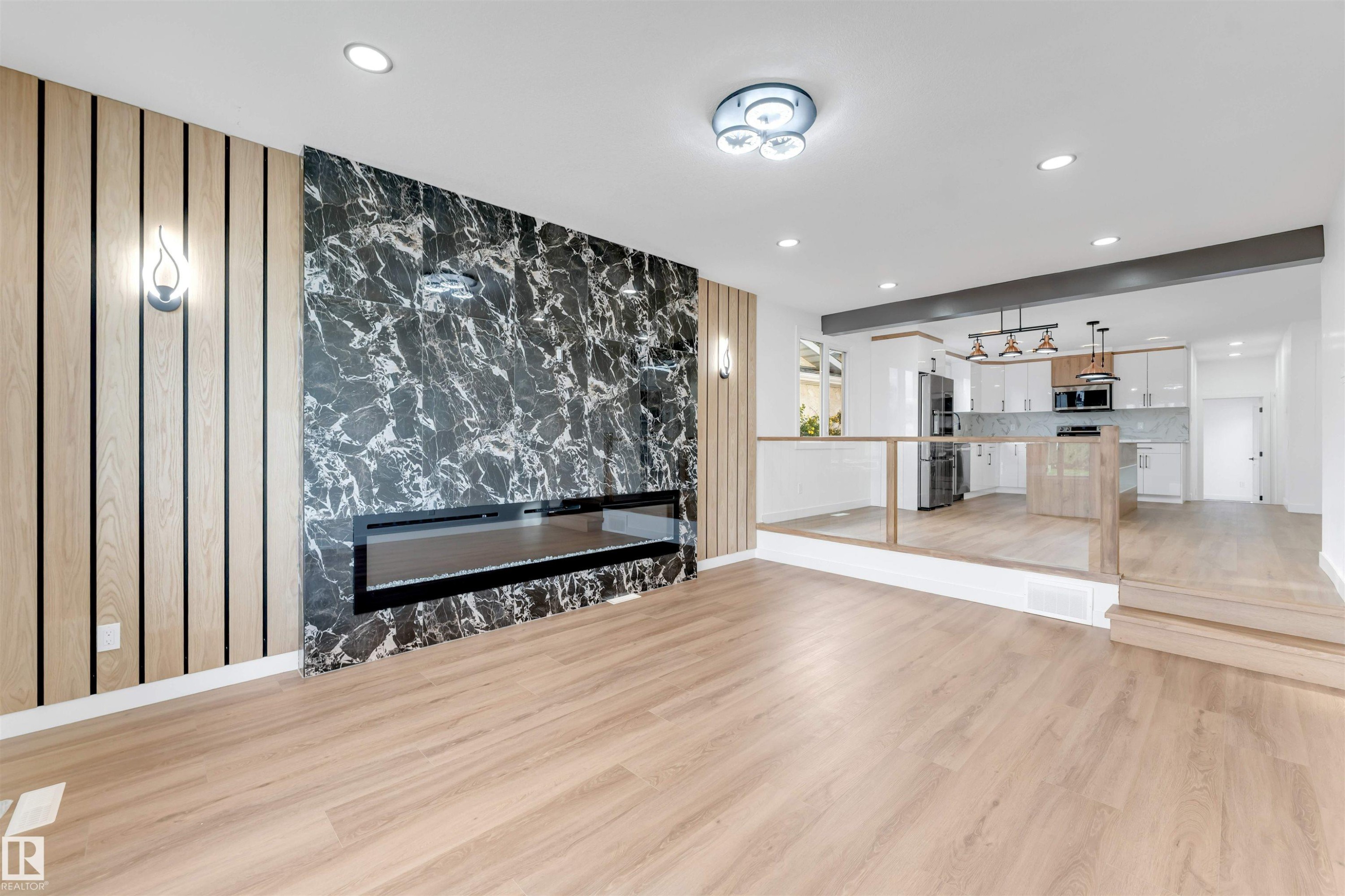 Spacious living area featuring wood-finish flooring, a modern linear fireplace with a black marble-patterned surround, and vertical wood slat wall accents - 5512 150 Avenue, Edmonton, AB
