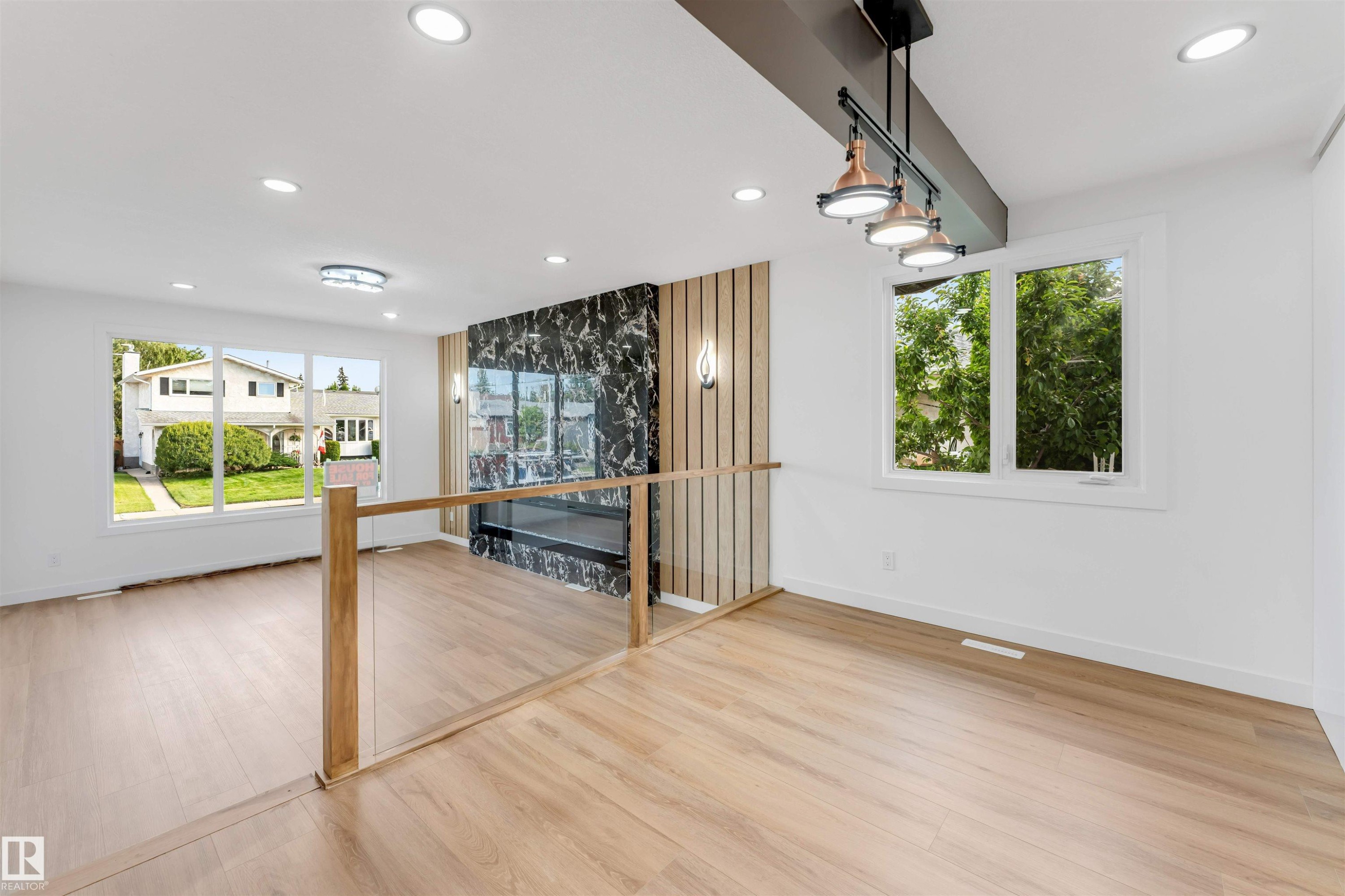Open-concept living area featuring light wood-finish flooring, a modern fireplace with a dark marbled surround, and a glass-paneled railing - 5512 150 Avenue, Edmonton, AB
