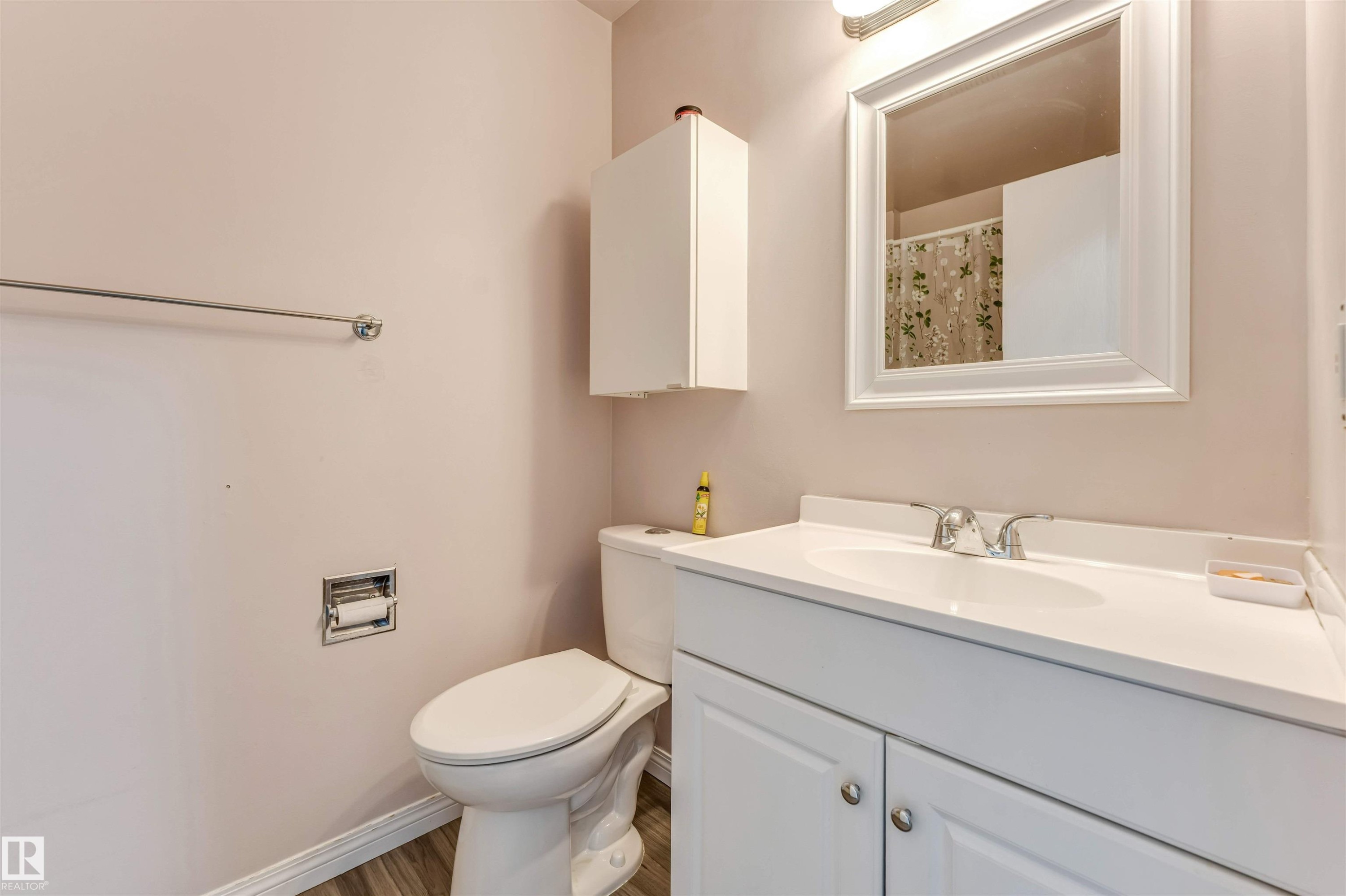 Bathroom featuring wood-finish flooring, a white vanity with an integrated sink and chrome faucet, a framed mirror, a wall-mounted cabinet, and a white toilet - 24 1651 46 Street, Edmonton, AB