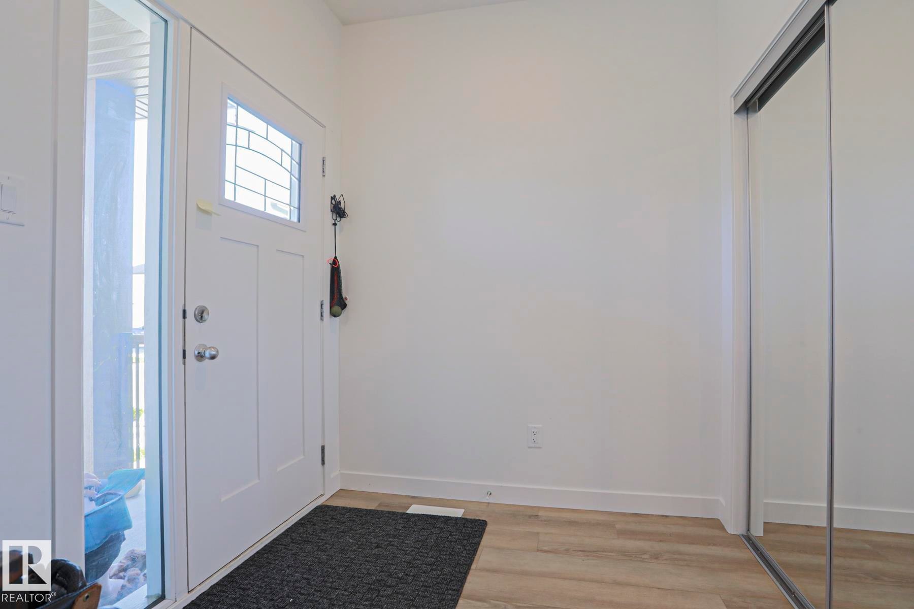 Entryway featuring a white paneled door with decorative glass inserts, a full-length sidelight, wood-finish flooring, and a mirrored sliding closet door - 33 Eton Link, Spruce Grove, AB