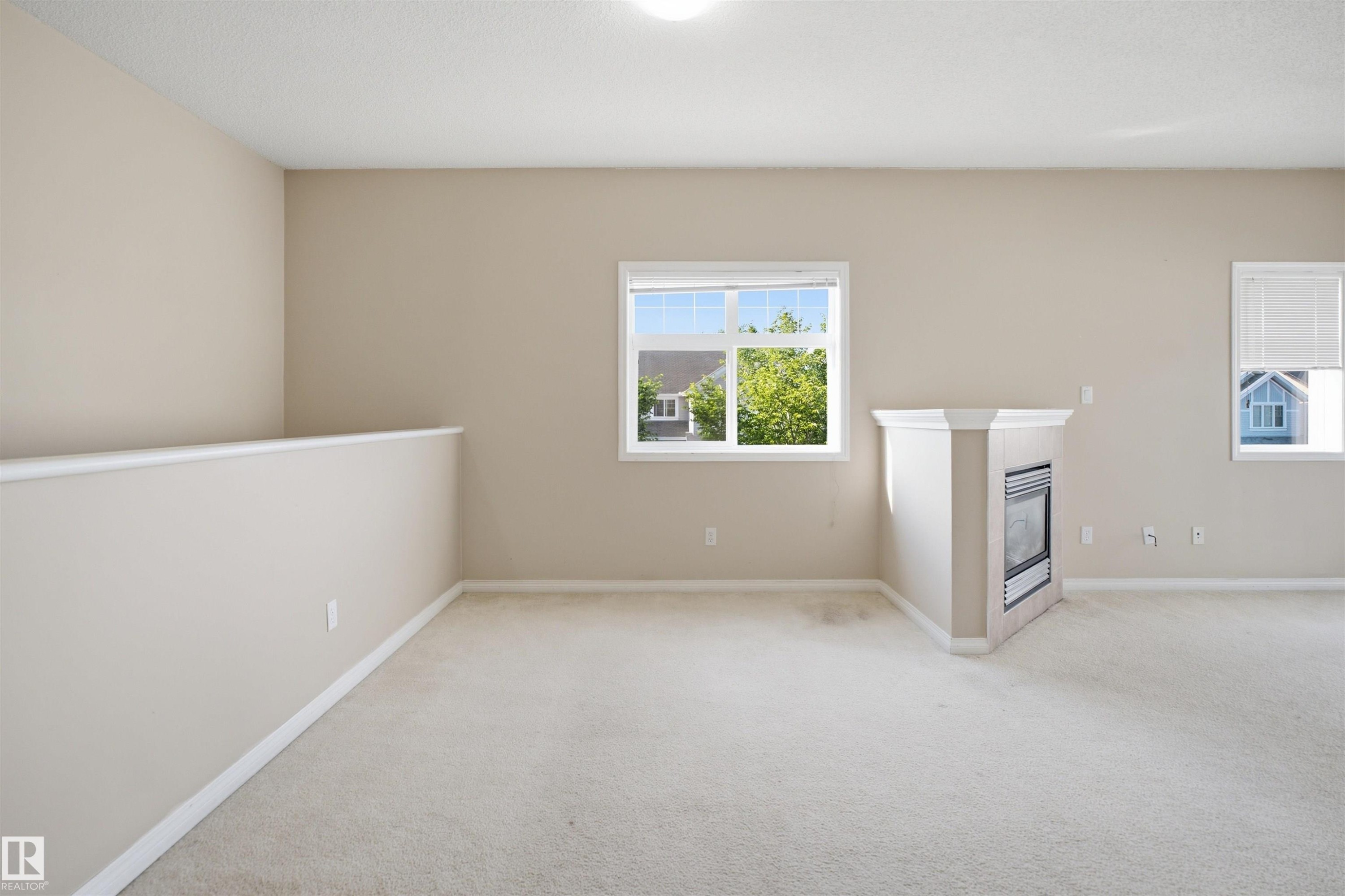 Carpeted room featuring a corner gas fireplace with a white mantel, multiple windows, and a partial wall with a white cap - 95 3040 Spence Wynd, Edmonton, AB