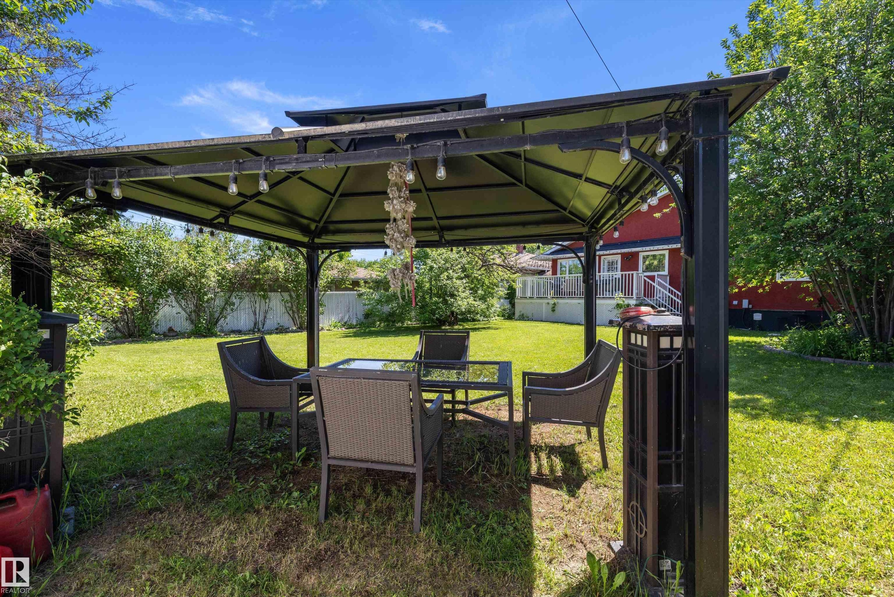 Shaded outdoor gazebo featuring string lighting, a manicured lawn, perimeter landscaping, and a distant red structure with a white deck - 11124 110A Avenue Nw, Edmonton, AB