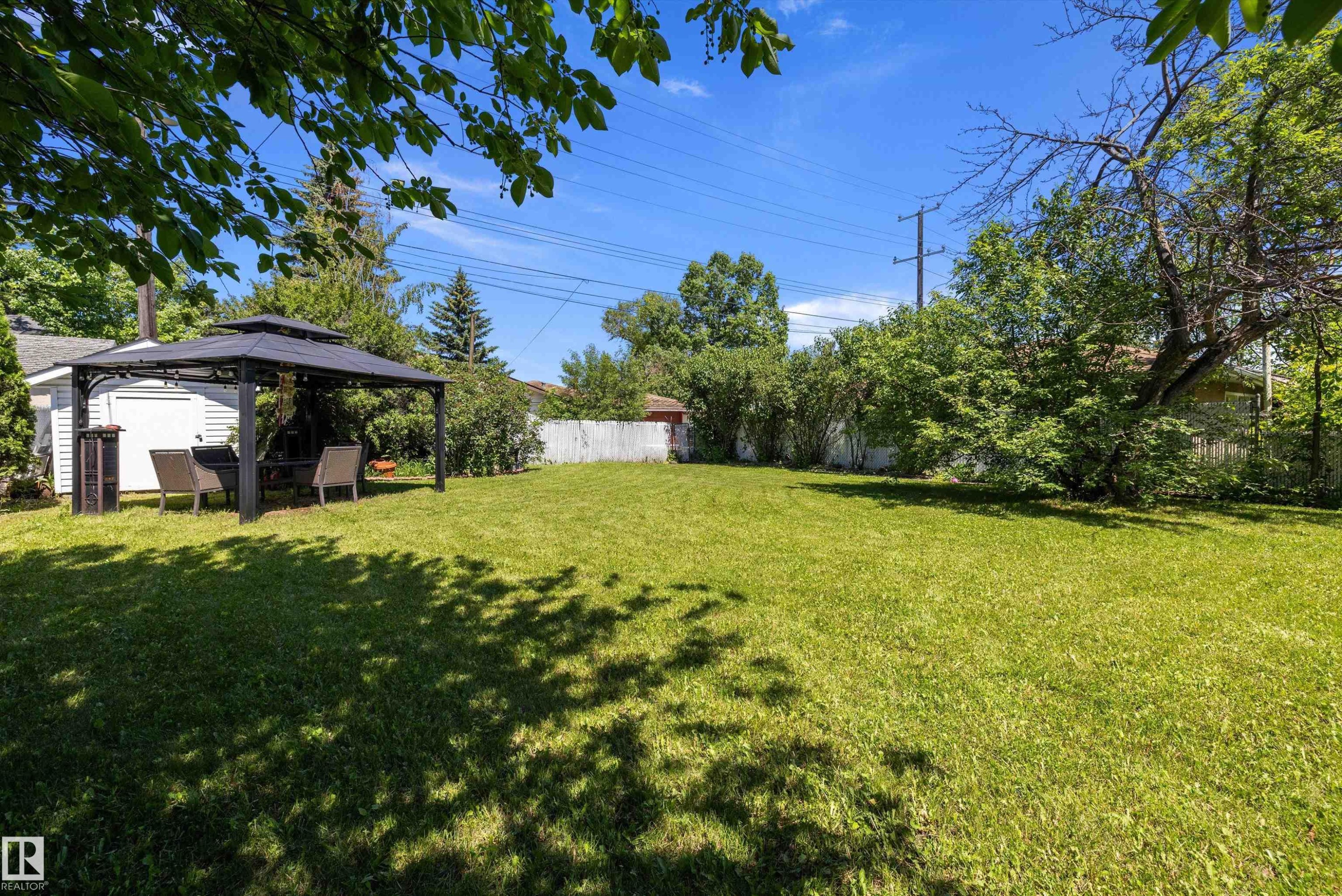 Spacious backyard featuring a large grassy area, mature trees, a white perimeter fence, a detached white structure, and a dark-toned gazebo - 11124 110A Avenue Nw, Edmonton, AB
