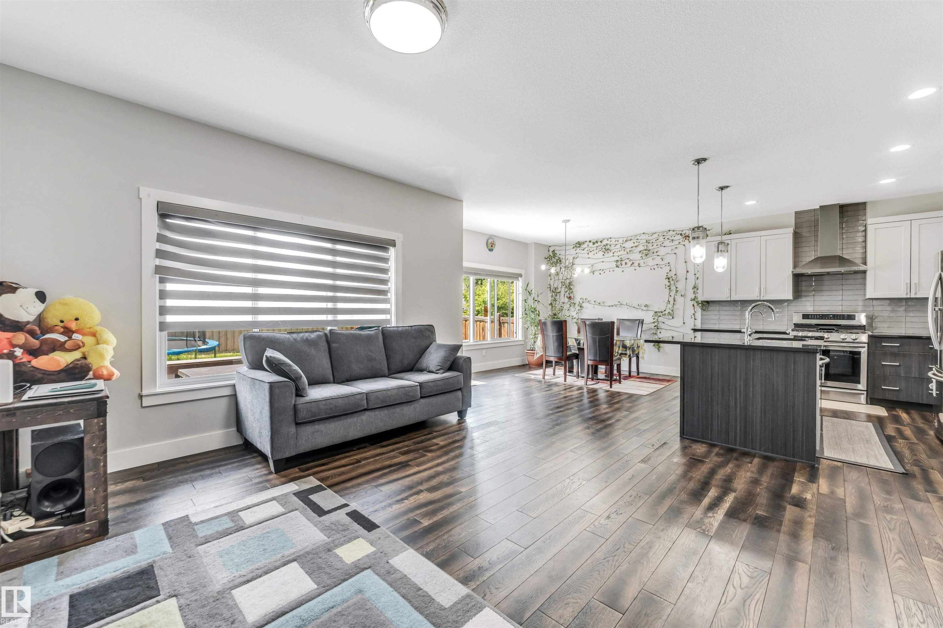 Open-concept living space featuring wood-finish flooring throughout, a modern kitchen with a large island and stainless steel appliances, and a dining area with pendant lighting - 8516 216 Street, Edmonton, AB
