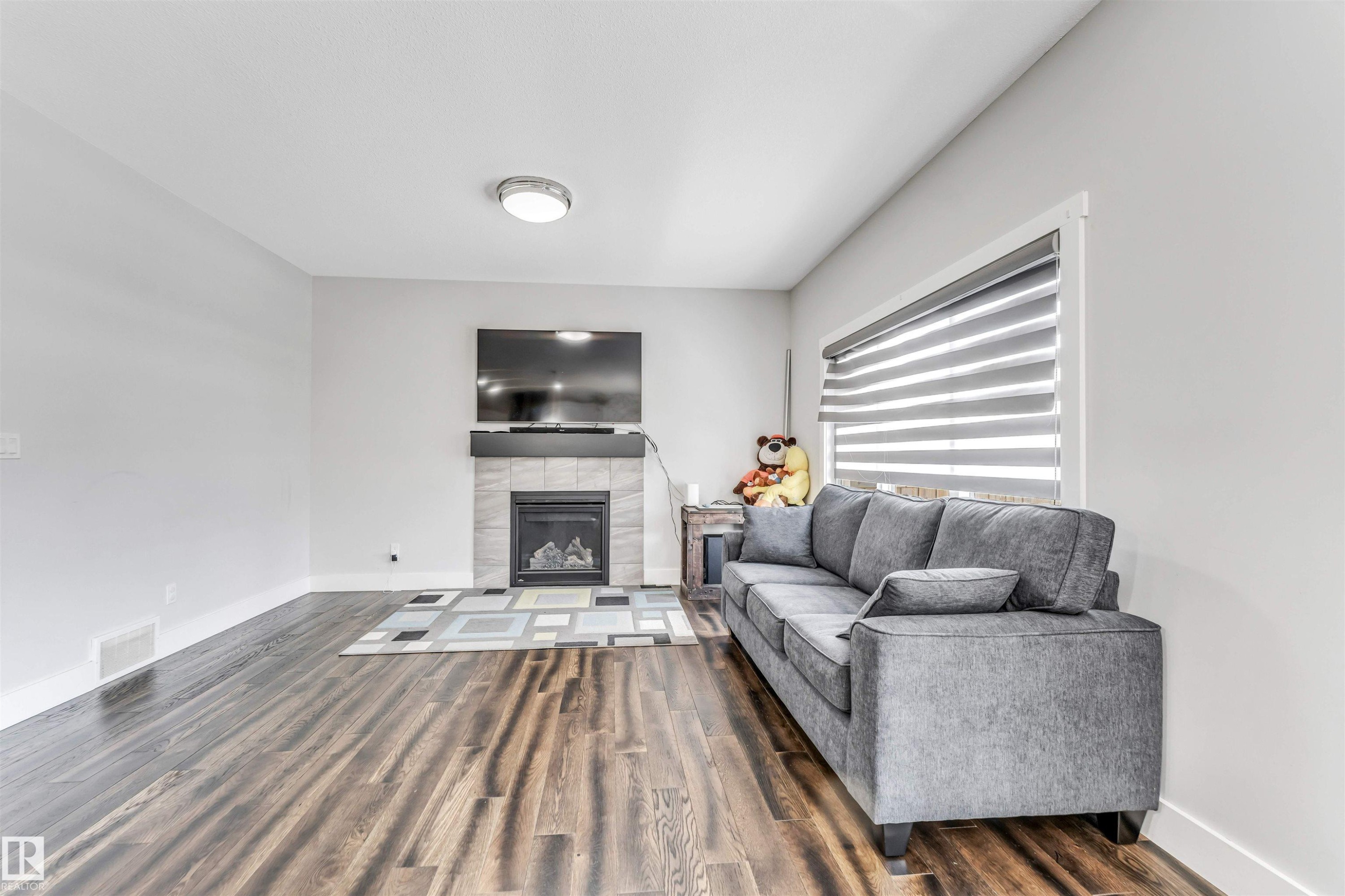 Spacious living area featuring wood-finish flooring, a tiled fireplace with mantel, and a large window with horizontal blinds - 8516 216 Street, Edmonton, AB