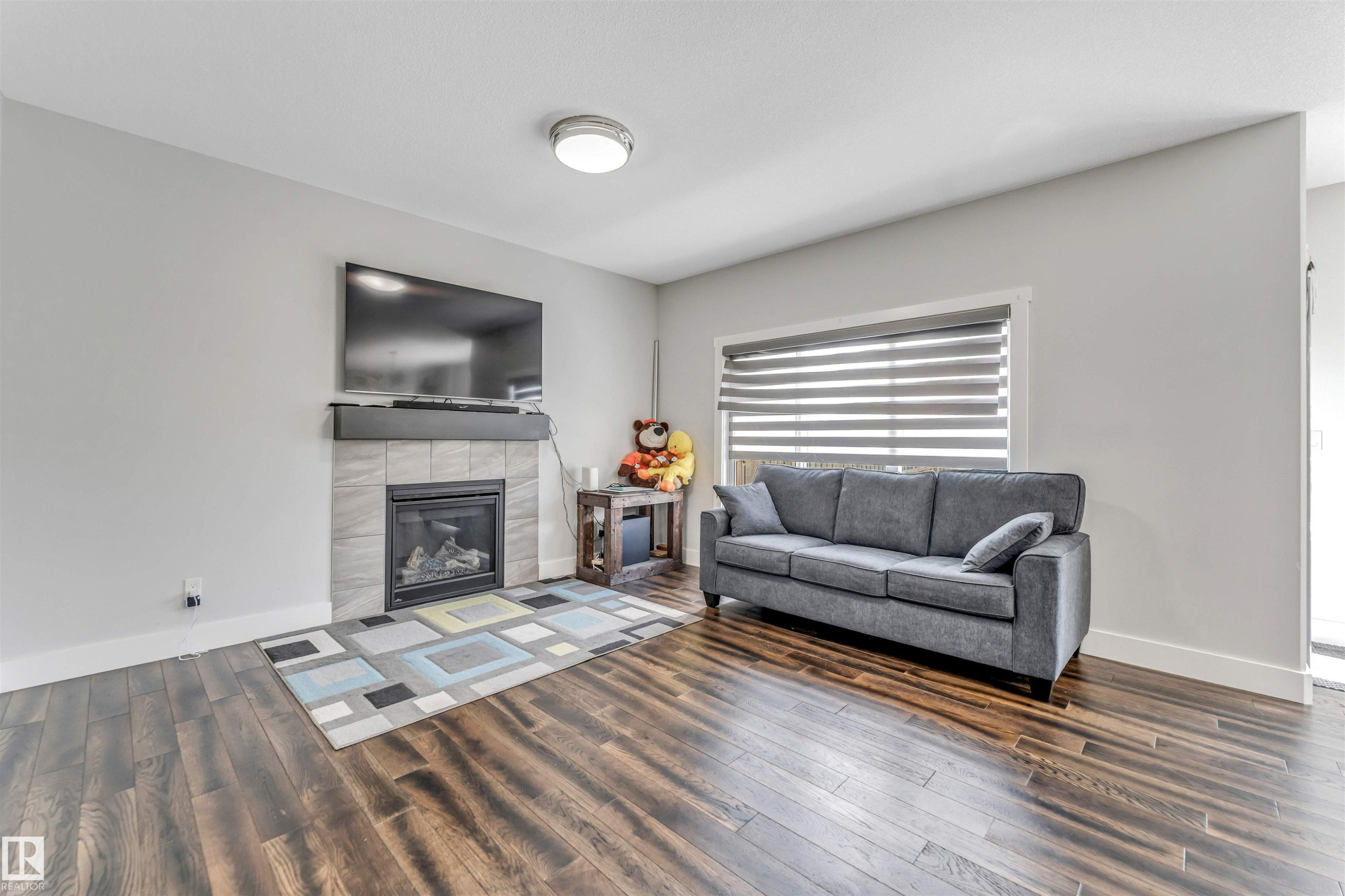 Living area featuring wood-finish flooring, a contemporary fireplace with a tile surround and dark mantel, a large window with horizontal blinds, and a flush-mount ceiling light fixture - 8516 216 Street, Edmonton, AB