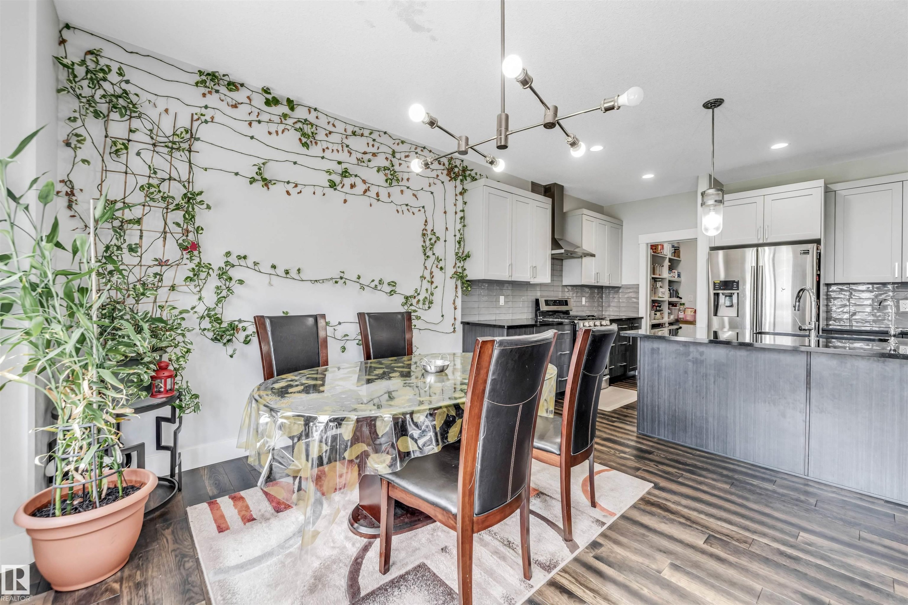 Modern open-concept space featuring wood-finish flooring, a contemporary linear chandelier, and a kitchen with stainless steel appliances, light upper cabinetry, and dark lower cabinetry - 8516 216 Street, Edmonton, AB