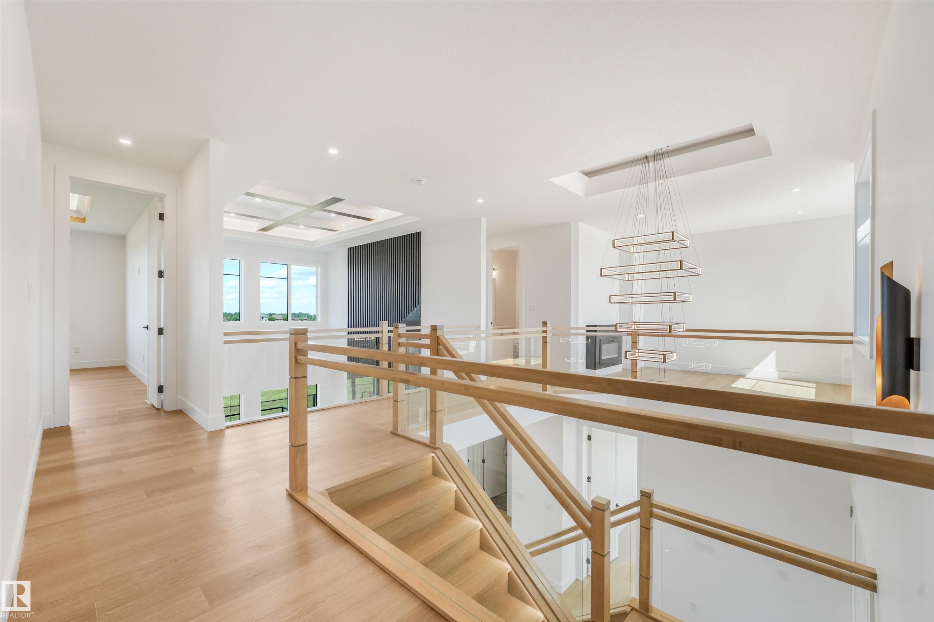 Upper-level landing featuring light wood-finish flooring, a contemporary chandelier, a glass and wood railing system, a tray ceiling with recessed lighting, and large windows - 1180 Genesis Lake Boulevard, Stony Plain, AB