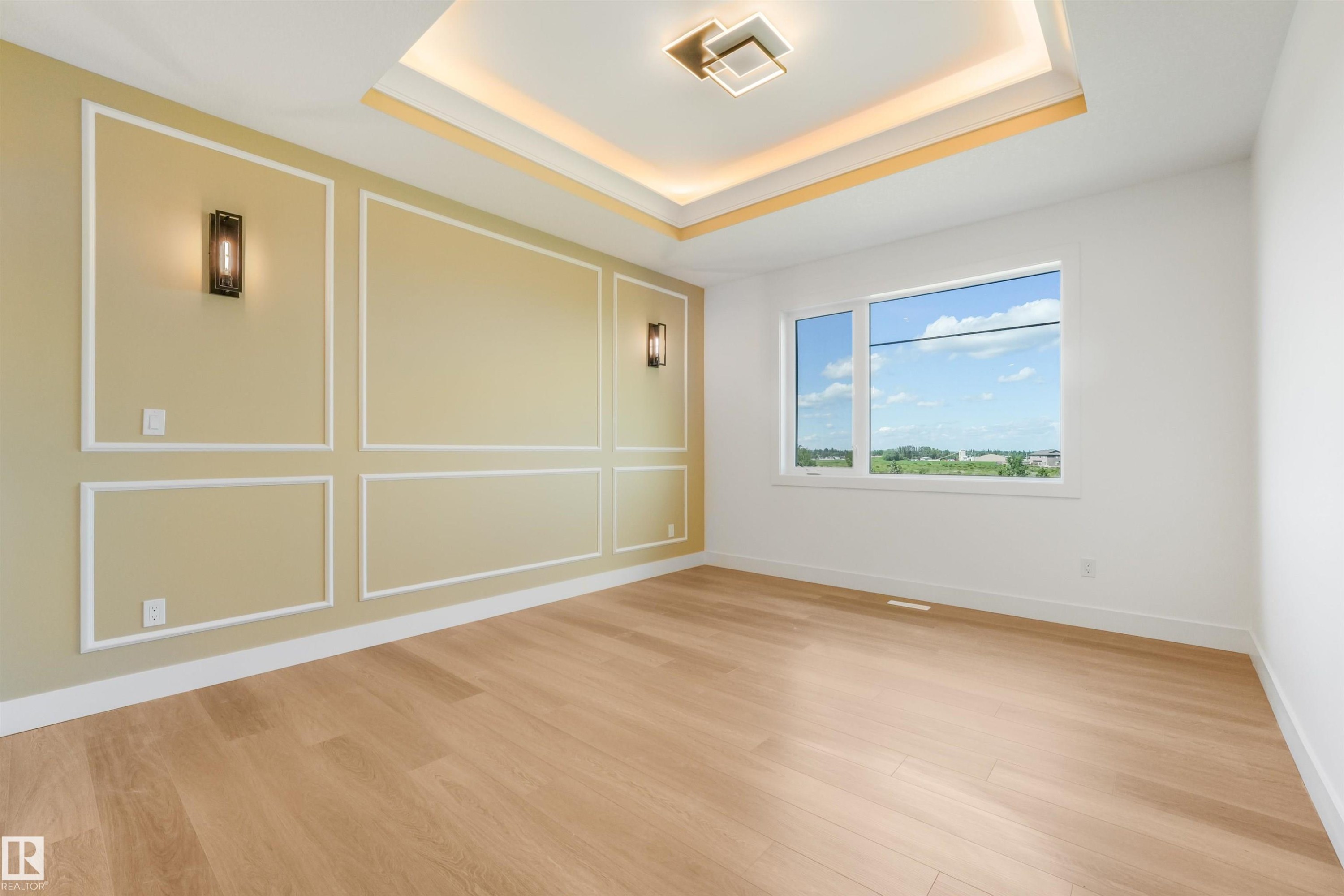Spacious room featuring light wood-finish flooring, a tray ceiling with recessed lighting and a central flush-mount fixture, a paneled accent wall with integrated sconces, and a large window with white trim - 1180 Genesis Lake Boulevard, Stony Plain, AB