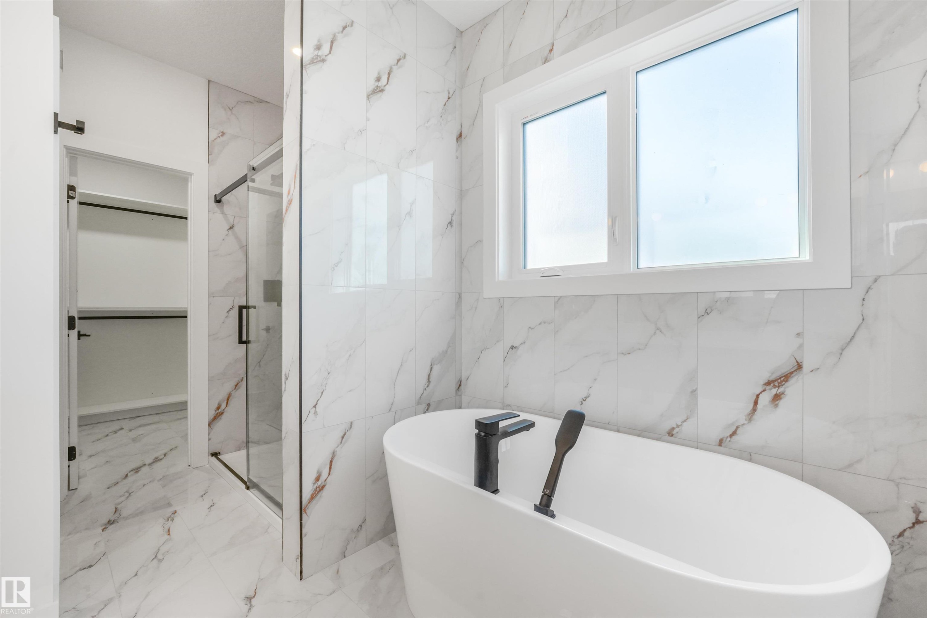 Freestanding oval bathtub with matte black floor-mounted faucet, surrounded by large format marble-look tile walls and flooring - 1180 Genesis Lake Boulevard, Stony Plain, AB