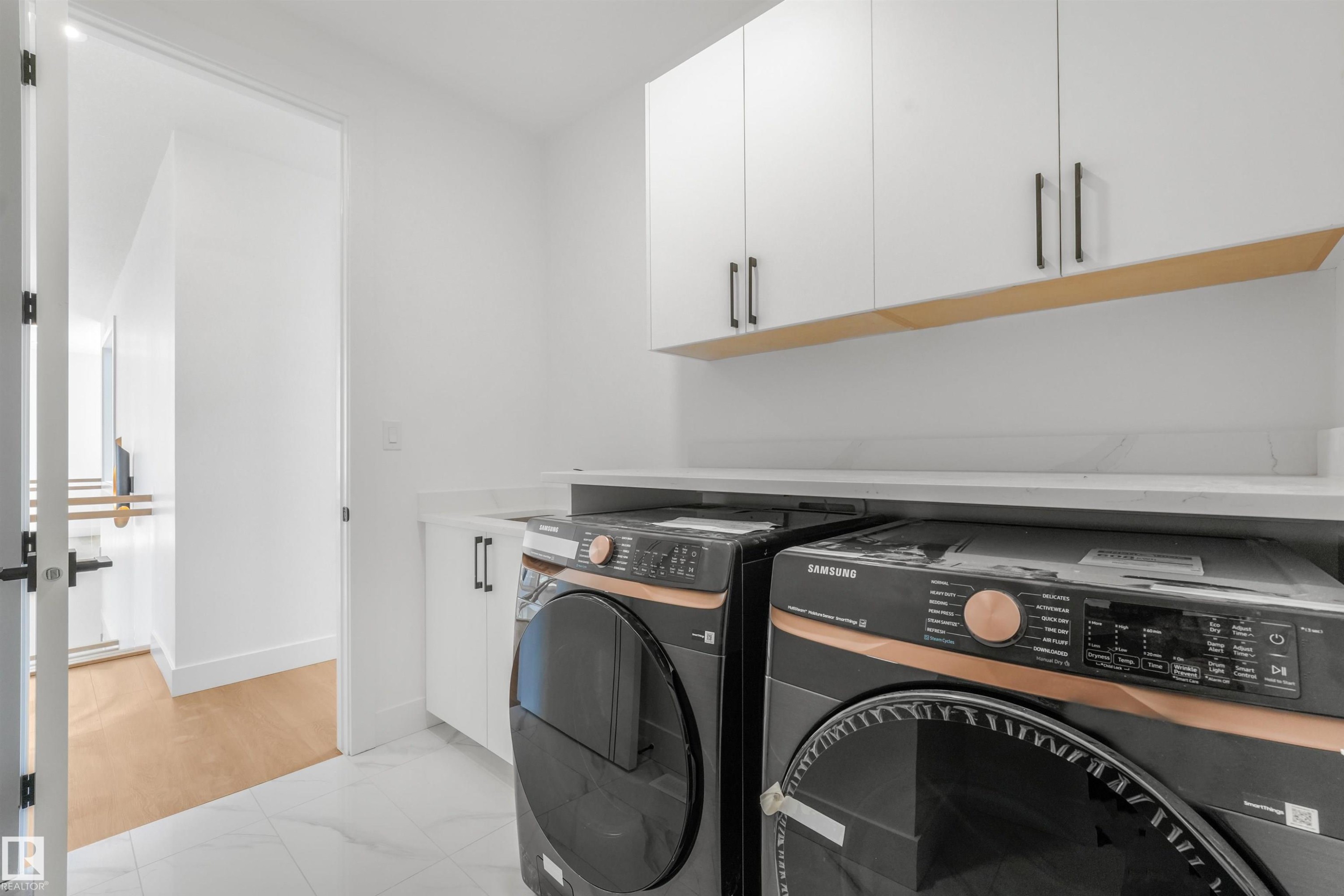 Dedicated laundry area featuring white upper cabinetry with matte black hardware, a wood-finish accent shelf, and a light-toned countertop - 1180 Genesis Lake Boulevard, Stony Plain, AB