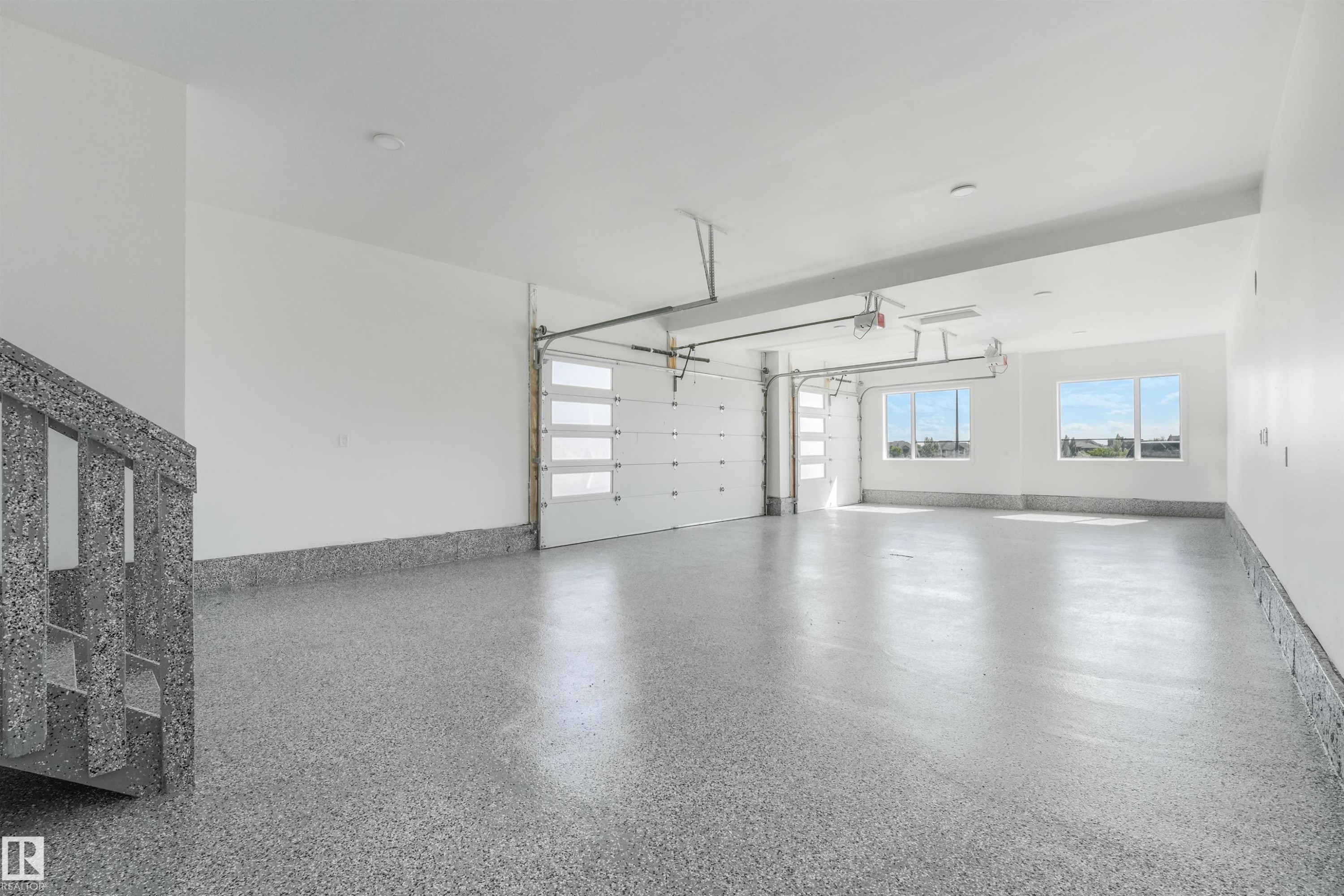 Spacious garage featuring a speckled epoxy floor finish, two modern garage doors with clear paneling, and ample natural light from multiple windows - 1180 Genesis Lake Boulevard, Stony Plain, AB