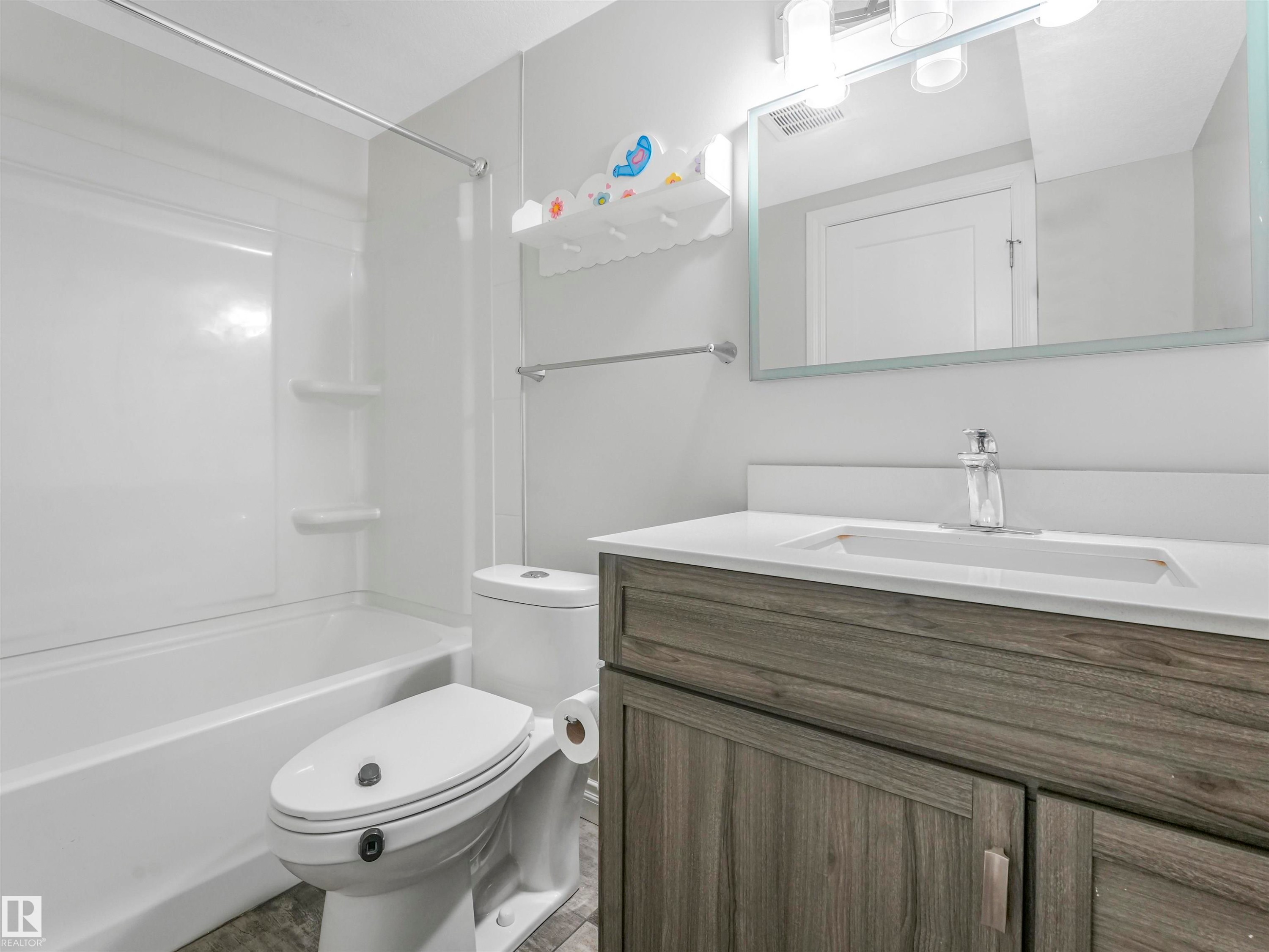 Contemporary bathroom featuring a white tub/shower combination with integrated shelving, a wood-finish vanity with a white countertop and integrated sink, a sleek wall-mounted mirror, and tile-style flooring - 20507 99 Avenue, Edmonton, AB - Indoor Photo Showing Bathroom